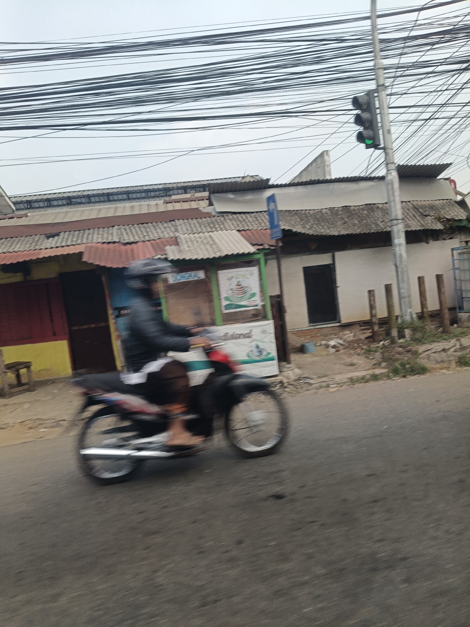 This is a motion-blurred, medium-angle, outdoor, daytime photograph of a man riding a motorcycle on a paved road. The man is wearing a black helmet and a black jacket over a white shirt. He is on a dark-colored motorcycle. The motorcycle's wheels are in motion. The road is gray and has some debris. To the left of the road is a building with a red and yellow facade and a corrugated metal roof. In front of the building are a small wooden stool and some signage. To the right of the road is another building with a white facade and a corrugated metal roof. A traffic light is visible on a pole to the right of the building, with a green light illuminated. Numerous power lines are strung across the top of the image. The overall environment suggests a street scene in a built-up area, possibly in Saruni, Indonesia, given the context.