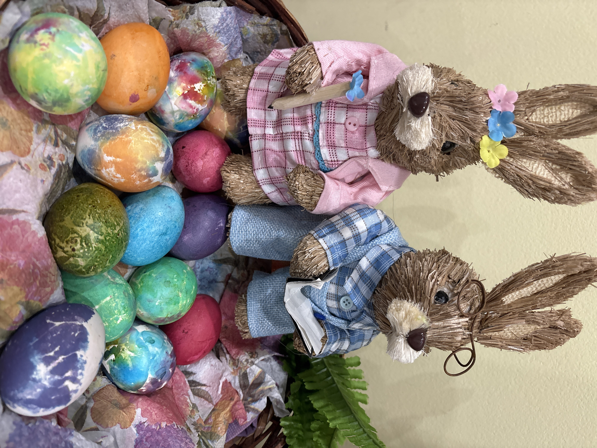 The image shows a basket filled with colorful Easter eggs and two straw bunny figurines. The Easter eggs are dyed in a variety of colors, including green, orange, blue, purple, and pink, with some having a marbled or swirled appearance. The eggs are arranged in a pile on top of a floral-patterned tissue paper inside a brown wicker basket.
Two straw bunny figurines are positioned next to the basket of eggs. One bunny wears a pink plaid shirt and a pink jacket, and has a small pencil behind the ear and flowers on its head. The other bunny wears a blue plaid shirt and has eyeglasses. The bunnies are lying down.
A green fern is visible near the bottom of the image, and a light yellow surface can be seen in the background. The location in Sofia, Bulgaria cannot be identified from the image.