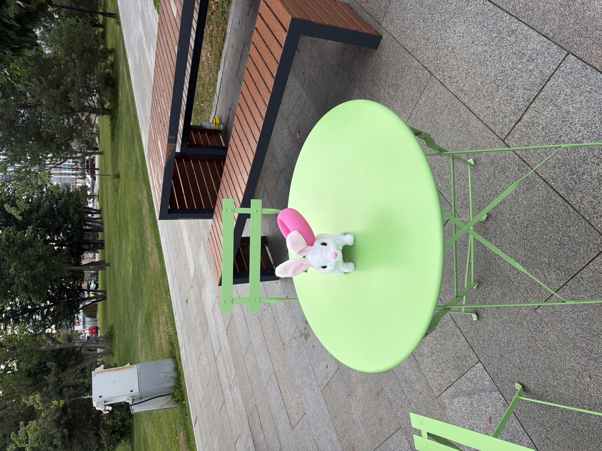 In an outdoor urban park setting, a small, white plush toy rabbit with pink ears and a pink collar is positioned on a bright green, round metal table. The table has a classic bistro design with foldable legs. Adjacent to the table, a matching green metal chair is visible. In the background, a long, brown wooden bench with a dark metal frame sits on a paved area. Beyond the seating area, a neatly manicured green lawn stretches out, bordered by trees and a pathway. A utility box with white and gray panels stands near the edge of the lawn. The overall scene depicts a quiet, possibly public, outdoor space.