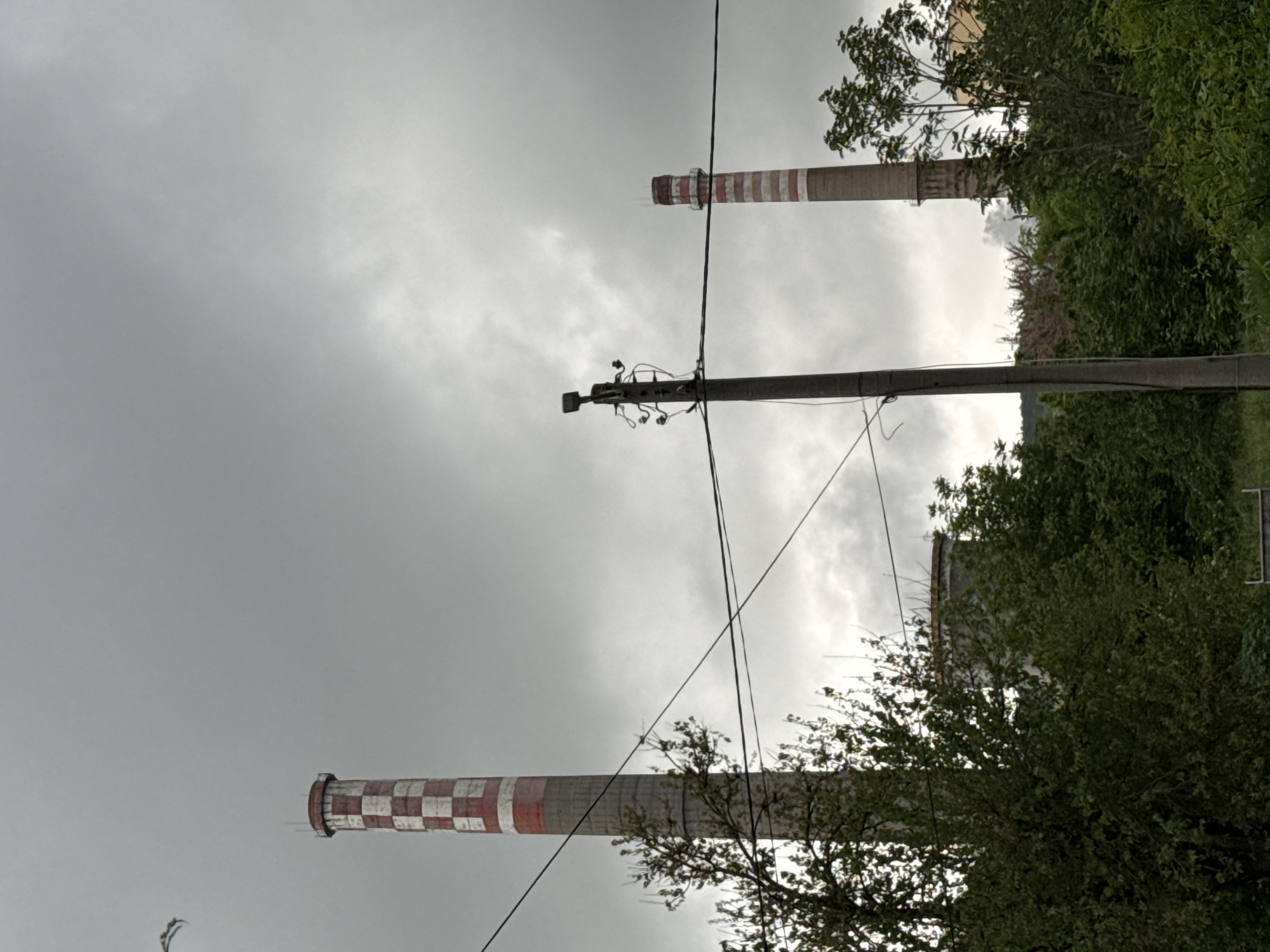 The photograph depicts a landscape in Pleven, Bulgaria, characterized by a cloudy sky and industrial elements. Visible are two tall smokestacks with alternating red and white bands. A utility pole with power lines stretches across the frame, intersecting the view of the smokestacks. The scene is bordered by trees and greenery. Buildings are partially obscured by vegetation. The photograph is taken from a low angle, emphasizing the height of the structures.
