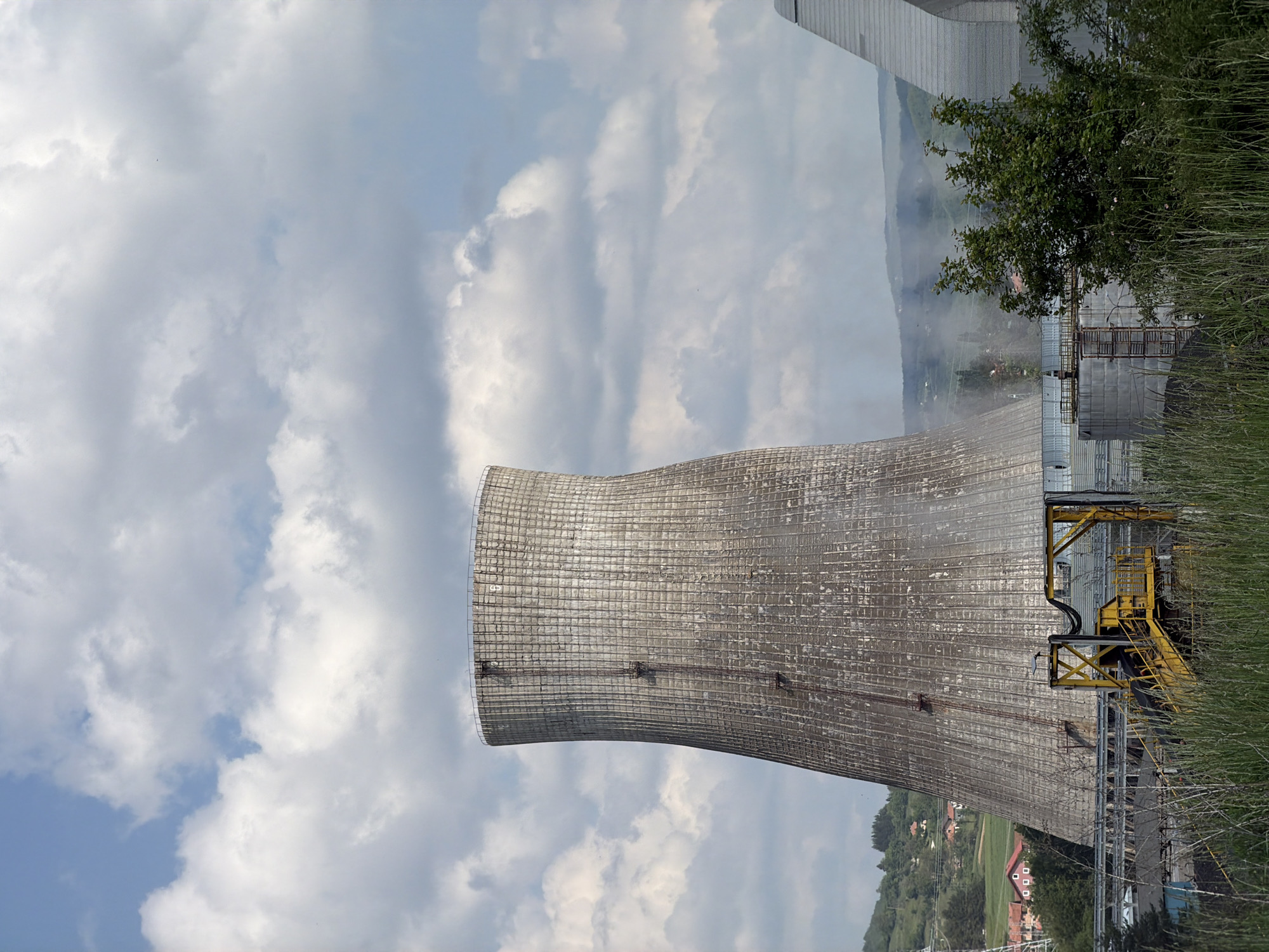 The image shows a cooling tower, likely part of a power plant or industrial facility. The tower is cylindrical with a flared top, constructed from concrete or a similar material. It is located in the city of Zabrđe, Montenegro.
The sky above the tower is filled with clouds, suggesting a partly cloudy day. To the side of the tower, there is some vegetation, including grass and trees. A structure with yellow supports can be seen adjacent to the tower. In the background, there are hills or mountains with some settlements visible. There is no apparent sign of any people or actions.