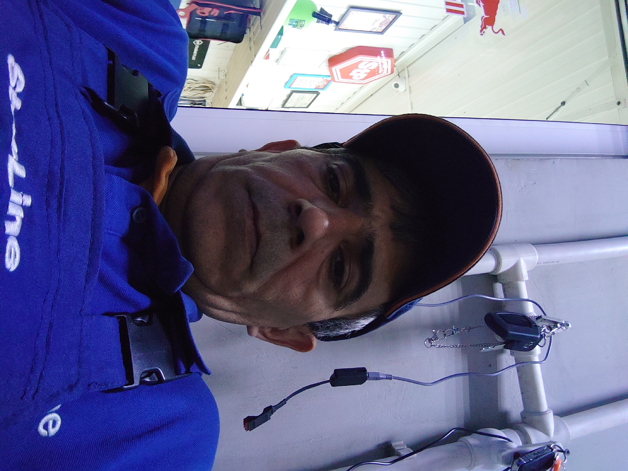 The image is a selfie taken by a middle-aged man wearing a blue work shirt and an orange-brimmed baseball cap. The man has short, graying hair and is looking directly at the camera with a neutral expression. He is wearing a blue shirt with black buckles on the front. In the background, there are white pipes, a key on a chain hanging from one of the pipes, and a red bull logo on the left side of the image. There are also shelves with various items, including a green spray bottle and a red hexagonal sign with "Stp" written on it. The setting appears to be a workshop or garage. The photo was taken in Belorechensk, Russia.