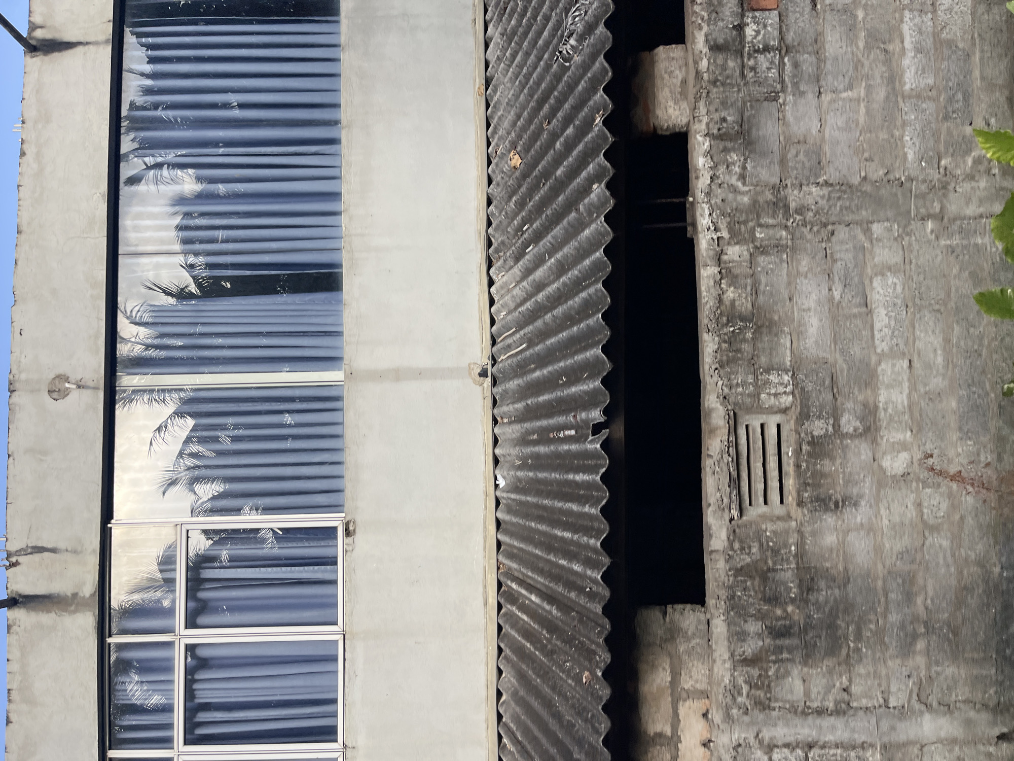 The image shows a close-up view of a two-story building, likely in Maggona, Sri Lanka, focusing on the facade. The upper story features a large window with blue curtains and a reflection of palm trees. The lower story has a corrugated metal roof supported by brickwork, and a rough concrete wall with a ventilation grille. The scene appears to be an exterior view, possibly a residential or commercial structure. There are no people or discernible actions visible in the image. The environment is a building exterior, with a clear sky visible in the top portion of the frame. The overall impression is one of a simple, functional building.