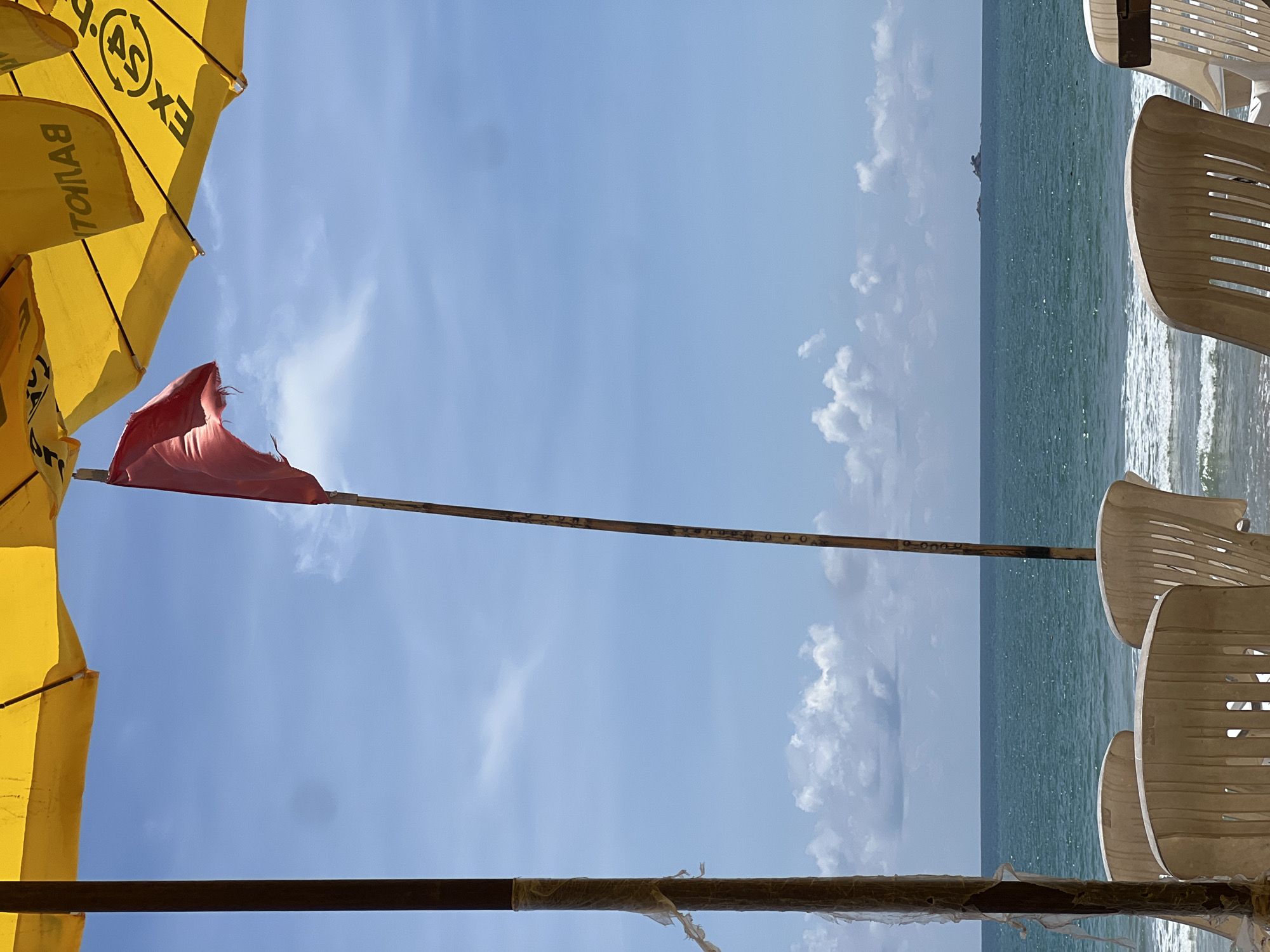 A red flag waves on a wooden pole in front of a bright blue sky with scattered white clouds, partially obscured by the yellow fabric of an open beach umbrella. To the left and right of the flag pole, brown wooden poles support the umbrella. The sea, a vibrant turquoise, stretches to the horizon, where a small cluster of rocks can be seen. In the foreground, several empty, weathered plastic beach chairs are arranged facing the sea. The scene is set on a beach, with waves gently lapping the shore. The location is identified as Unknown, Unknown.