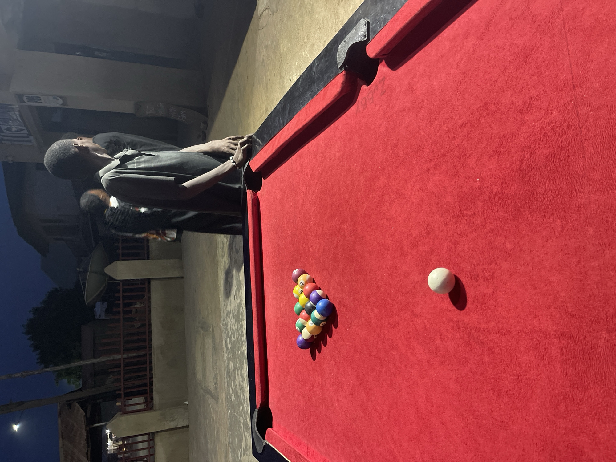 The image presents a nighttime scene in Unguwar Gabas, Nigeria, centered around a red-felted pool table. On the table, a full rack of 15 numbered pool balls, including solids and stripes of various colors, is neatly arranged. A single white cue ball rests separately on the felt.
To the left of the table, a man in a dark polo shirt and trousers stands, his hands resting on the table's edge. He wears a bracelet on his left wrist and appears to be looking off into the distance or at something outside the frame. Another individual is partially visible behind him, also dressed in dark attire.
The setting is dimly lit, indicating it is nighttime, with artificial light sources creating distinct shadows from the pool balls. The background reveals an outdoor or open-air environment with concrete walls, metal railings, and a prominent satellite dish. A tree is visible under a dark sky, with a distant light source, possibly a street light or the moon, casting a soft glow. The overall atmosphere suggests a casual, relaxed gathering where a game of pool is either about to commence or is currently paused. Faint and illegible text can be observed on a wall in the distant background.