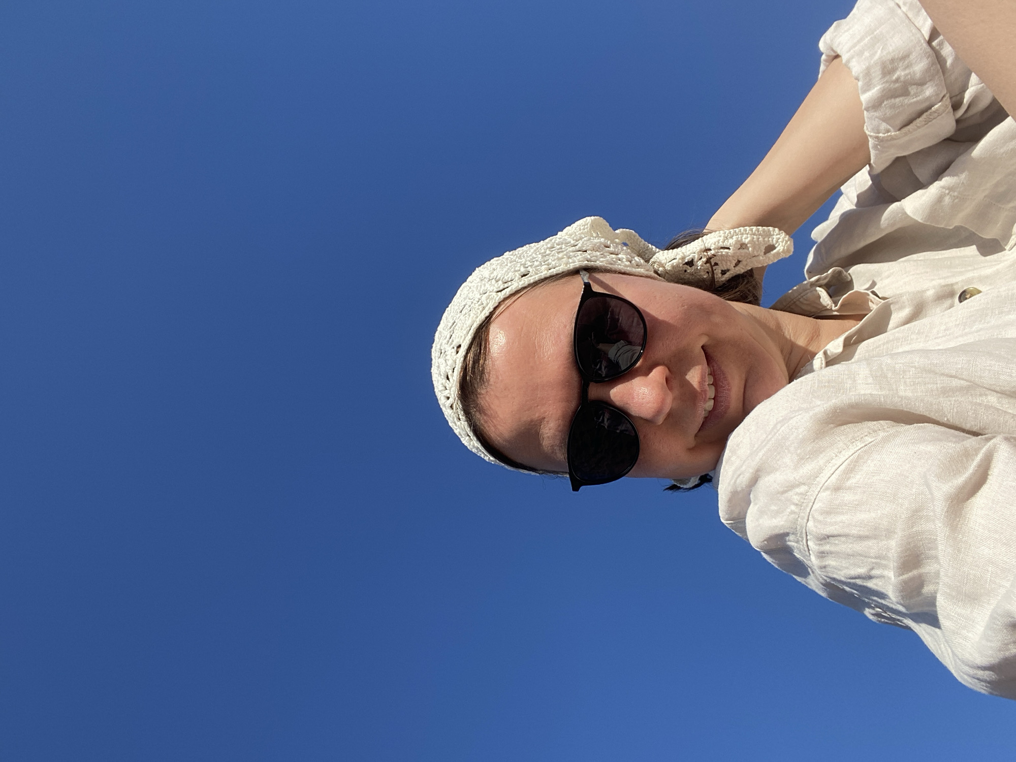 A woman wearing a white crocheted headscarf and dark sunglasses is angled against a clear blue sky. She is also wearing a light-colored, button-down shirt. The image appears to have been taken from a low angle, looking up at the subject. The exact location in Chaikovsky, Russia, cannot be determined from the image alone.