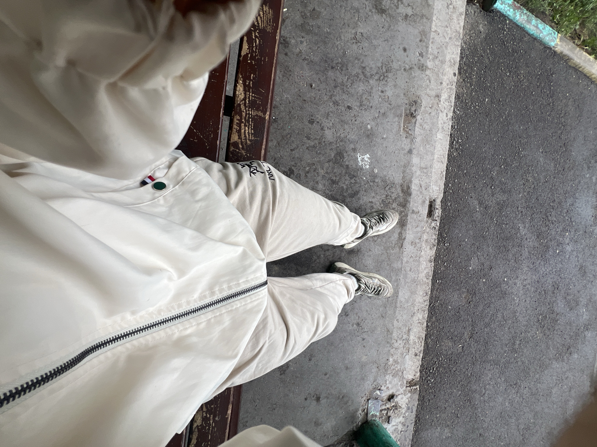 A first-person perspective shot shows a person sitting on a wooden bench with their legs extended forward. The person is wearing light-colored sweatpants with black lettering on the left leg and white sneakers with black laces. A light-colored jacket with a black zipper is visible in the foreground. The bench is situated on a concrete walkway next to a paved area with some grass and a curb painted in turquoise. The background is a dark asphalt surface. The scene appears to be outdoors, possibly in a park or along a street in Qarağandı city administration, Kazakhstan.
