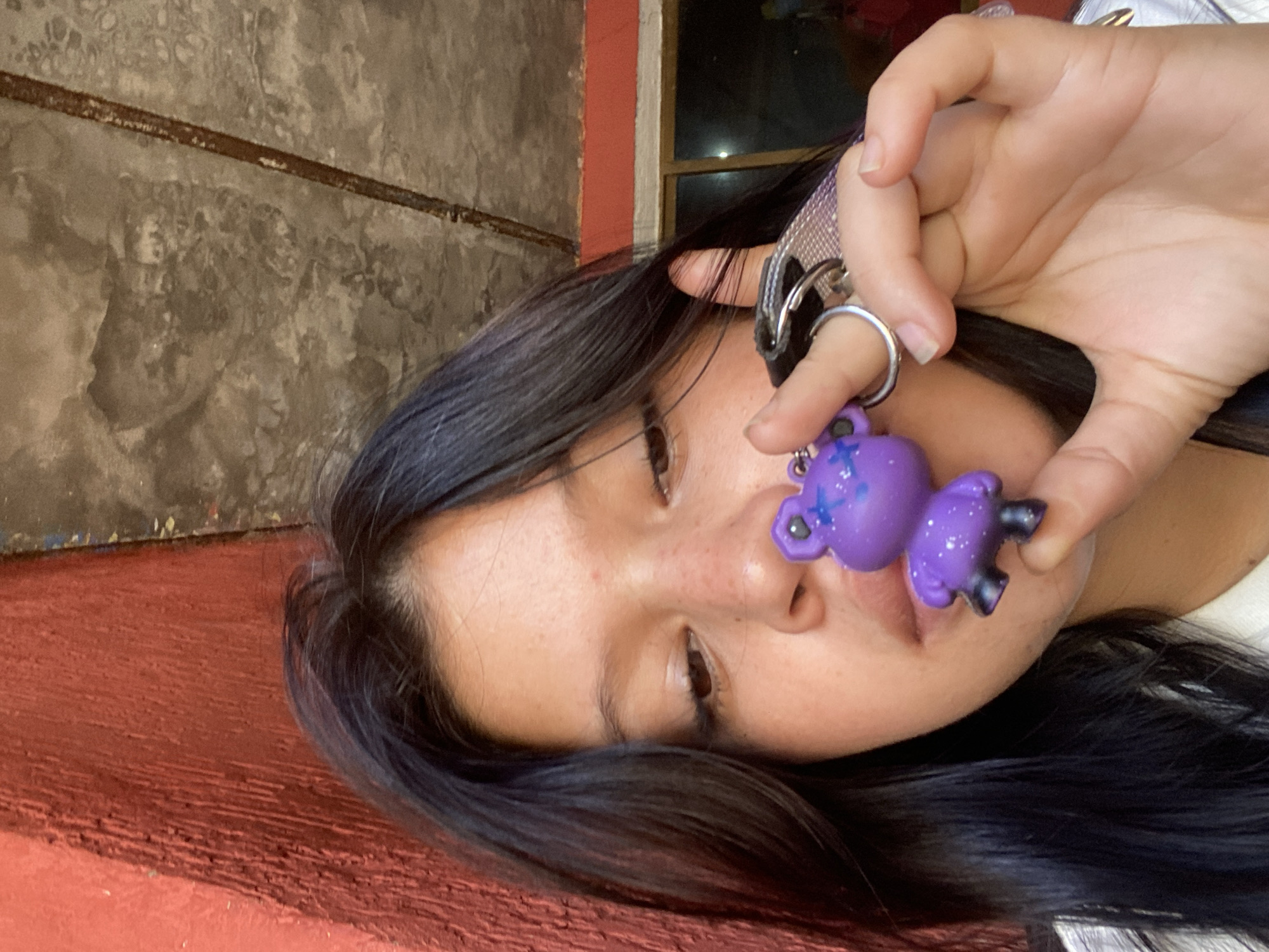 A young adult female is holding a purple plastic bear keychain in front of her face. She is lying on a textured red surface, presumably a ledge or bench. The background includes a grey concrete wall with a rusty horizontal band and a partially visible window frame with a red border. The location is not discernible from the image, but information provided specifies Juan Griego, Venezuela.