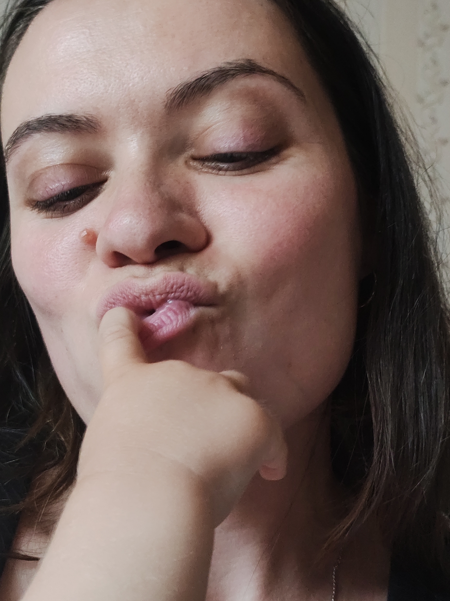 A close-up shot of a woman with fair skin and dark hair. She has her eyes mostly closed and pursed lips, with a finger touching her lower lip. A mole is visible on the side of her nose. No location is identifiable.