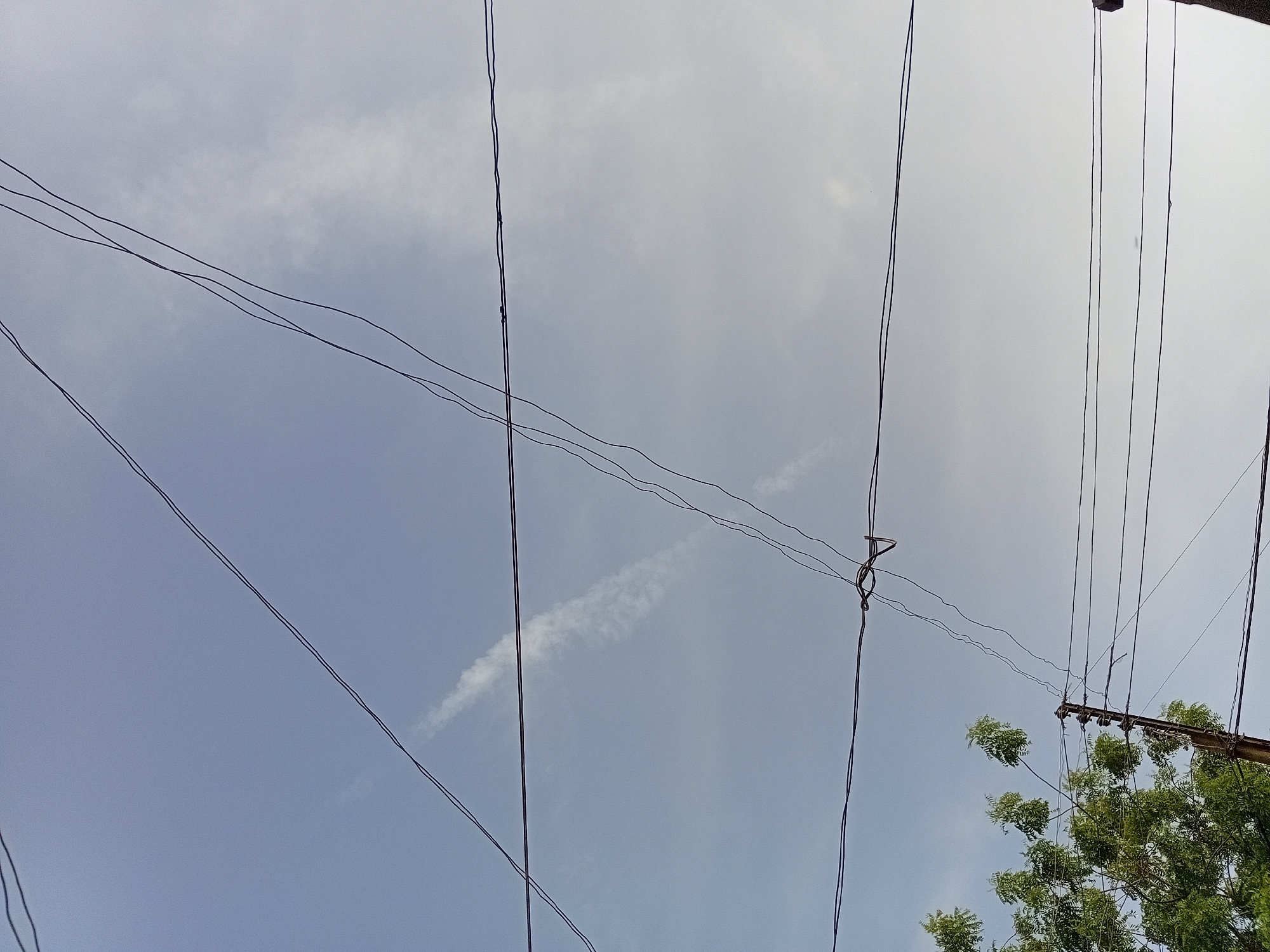 The image captures an upward-looking perspective of an intricate network of utility wires set against a partly cloudy daytime sky in Dakata, Nigeria. Numerous dark, thin wires crisscross the frame, creating a dense pattern. Some wires traverse diagonally from the upper left, while others descend vertically and horizontally across the central and right portions of the sky.
Towards the right side of the image, the top section of a utility pole is visible, supporting several wires via insulators. Below the pole and wires, the vibrant green foliage of a tree, likely with small leaves, occupies the bottom right corner, its branches reaching upwards.
The sky exhibits a mix of pale blue and soft grey-white clouds, indicative of partially overcast conditions. A distinct, elongated, wispy white cloud or contrail stretches horizontally across the mid-left area of the sky. No specific human activity or identifiable text is visible in the frame. The scene presents a common urban or semi-urban landscape, highlighting the overhead infrastructure for power or communication lines.
