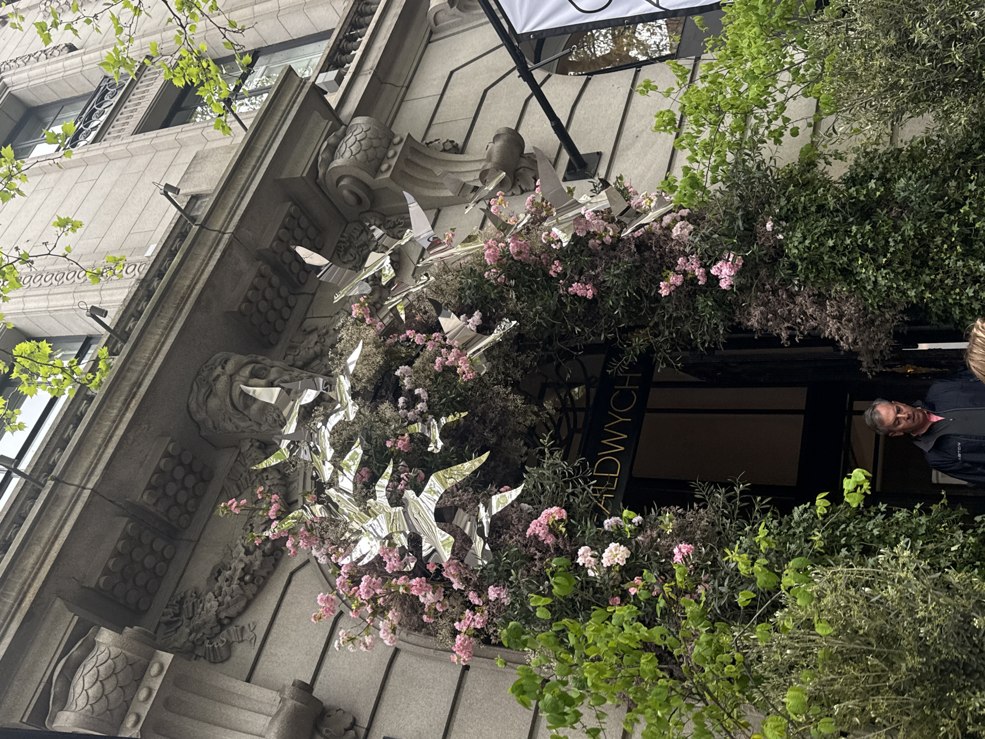 The photograph shows an exterior architectural detail in London, United Kingdom, likely at the Aldwych Theatre entrance. The building has an ornate facade, featuring carved details and mouldings. A floral arrangement adorns the entrance, intertwined with reflective, stylized bird figures. The flowers are primarily pink. Lush green foliage and shrubbery surround the entryway. A partial view of a man is visible to the right of the frame, possibly a patron or employee. The sign to the right of the entrance is black with "ALDWYCH" in gold lettering written vertically.
