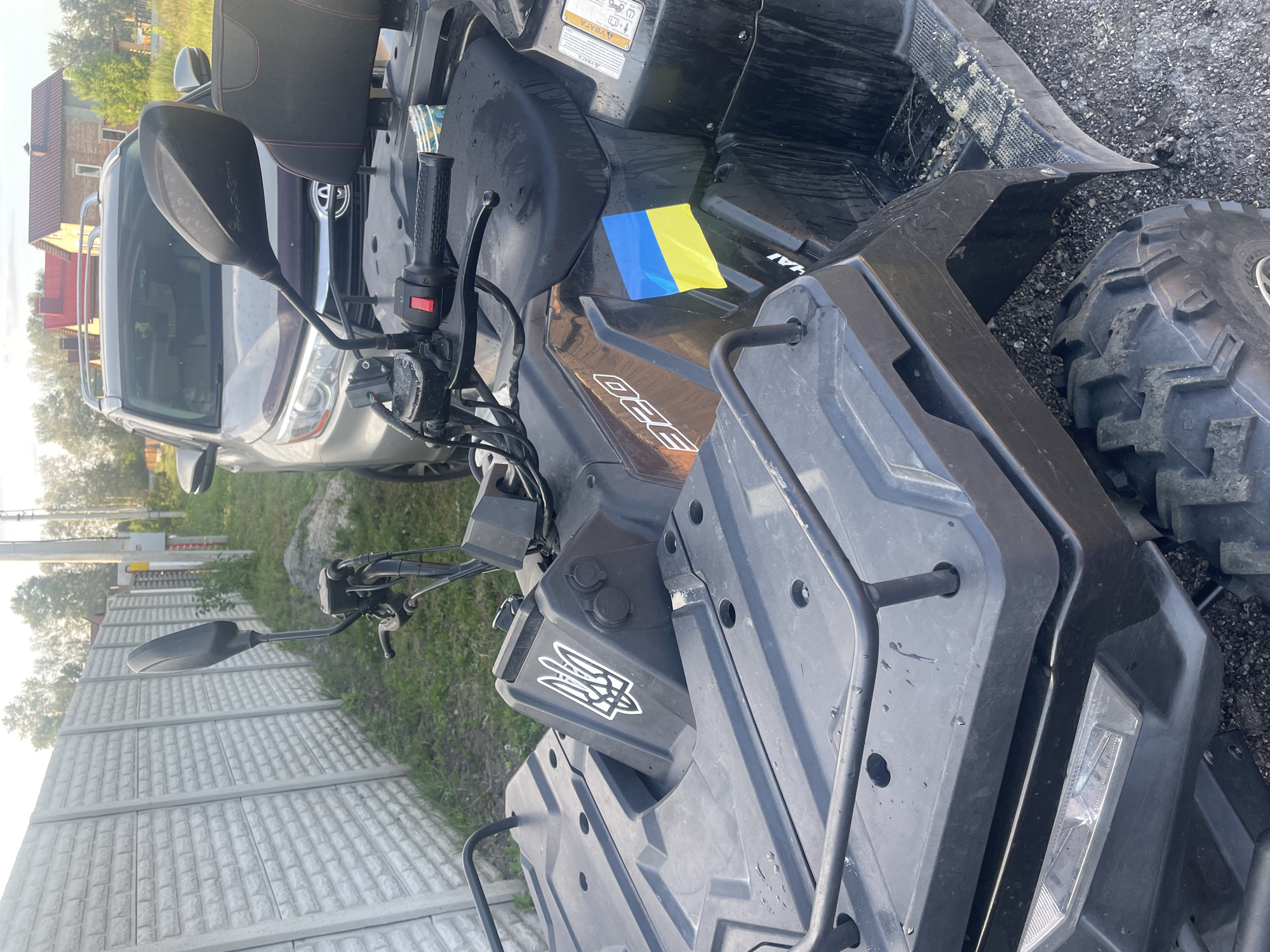 The image depicts an outdoor scene in Petrivka, Ukraine, featuring a black all-terrain vehicle (ATV) in the foreground. The ATV has a sticker of the Ukrainian Trident (Tryzub) emblem on its front body panel and a rectangular blue and yellow Ukrainian flag sticker on its side, near where the number "320" is also visible. The ATV's handlebars, rearview mirror, black textured seat, a portion of its front storage rack, and a knobby tire are discernible. The vehicle appears somewhat damp and shows signs of dirt.
In the mid-ground, a silver or light grey Toyota Highlander SUV is parked. The front grille, headlights, and roof rails of the SUV are visible.
To the left of the ATV, a long, light grey concrete block wall, featuring a brick-like texture, extends into the background. Sparse green grass and weeds grow at the base of this wall.
In the background, beyond the SUV and wall, numerous trees with green foliage are present. Several power poles and overhead lines are also visible. Two houses with reddish-brown roofs can be seen in the distance on the right side of the image, suggesting a residential or rural environment.
The ground where the ATV and SUV are located is covered with gravel and dirt. The lighting indicates either early morning or late afternoon with clear skies. No individuals are present in the image.