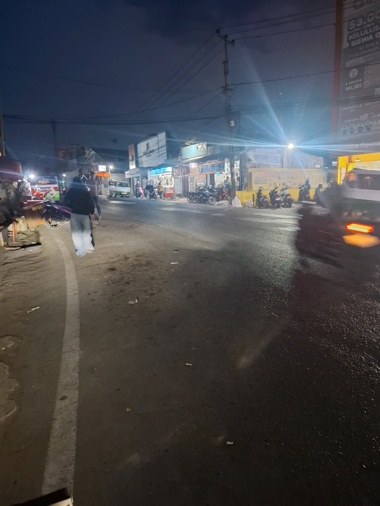 This is a street scene at night in Cibadak, Indonesia. A person in light-colored pants and a dark jacket walks away from the camera on the left side of a wet asphalt road. They are near a sidewalk with several parked motorcycles. Across the street, several shops are open with bright lights illuminating the area. A white van is parked on the right side of the road, its brake lights illuminated. Power lines are visible crisscrossing the dark sky overhead.