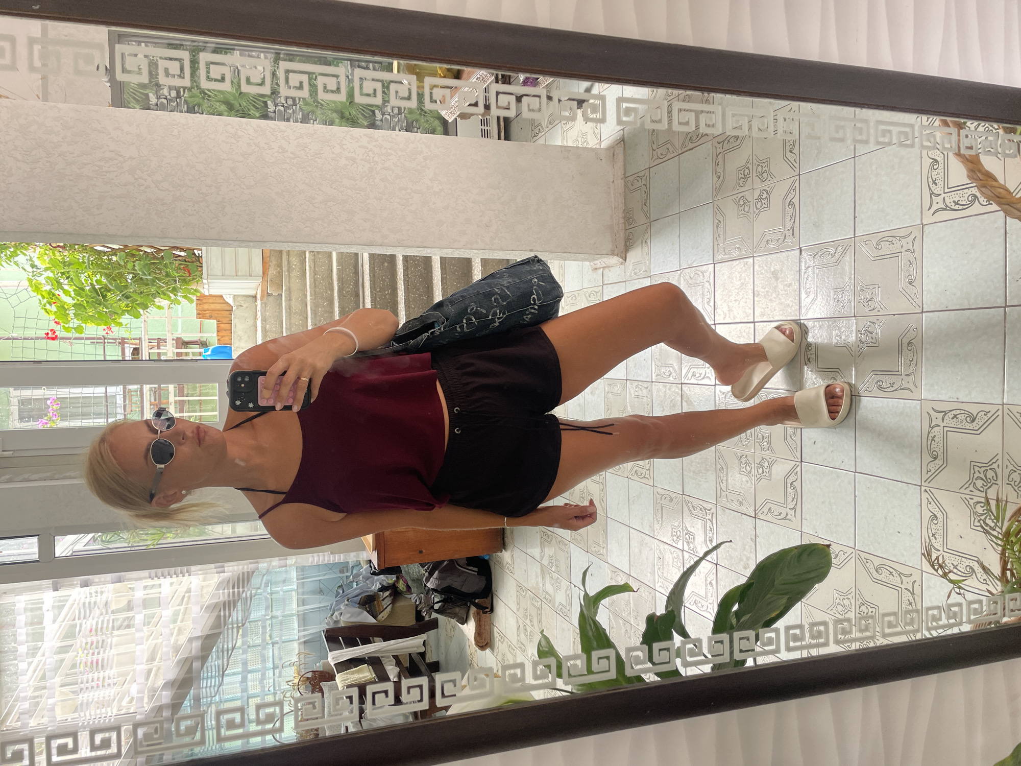 A woman with blonde hair pulled back stands in front of a full-length mirror, taking a selfie. She is wearing a dark red tank top, black shorts, and light-colored slide sandals. She has on round sunglasses and is holding her smartphone in her right hand. A dark denim bag with white lettering is slung over her left shoulder. The reflection in the mirror shows a tiled floor and a staircase leading up to an open doorway with greenery visible outside. In the background, through a window, a glimpse of the city of Gagra, Abkhazia is visible, characterized by its lush vegetation and Mediterranean-style architecture. The room appears to be an interior space with a tiled floor and patterned walls, possibly a home or a hotel room.
