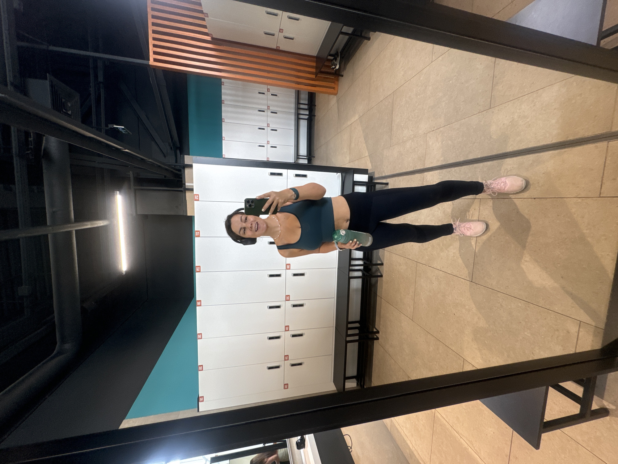 A young woman in workout clothes is taking a mirror selfie in a gym locker room. She is wearing a teal sports bra, black leggings, and pink athletic shoes. She has on headphones and is holding a green water bottle and a smartphone. The locker room has white lockers, black benches, and beige tiled flooring. The reflection shows more lockers and a wooden partition. The specific location in the city of Krasnoyarsk, Russia cannot be determined from the image.