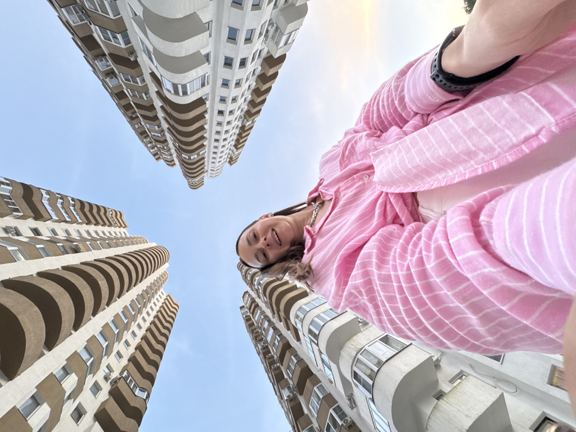 In an extreme low-angle, wide-angle shot, a woman wearing a pink striped shirt and a silver necklace smiles at the camera while standing against a backdrop of tall apartment buildings in Sochi, Russia. The buildings, with their curved balconies and white and tan facades, appear to tower over her, creating a sense of scale. The sky is a clear, pale blue, with a hint of sunset glow on the right side of the frame. The woman's face is well-lit, and her expression is relaxed and happy. The focus is primarily on the woman and the architecture.