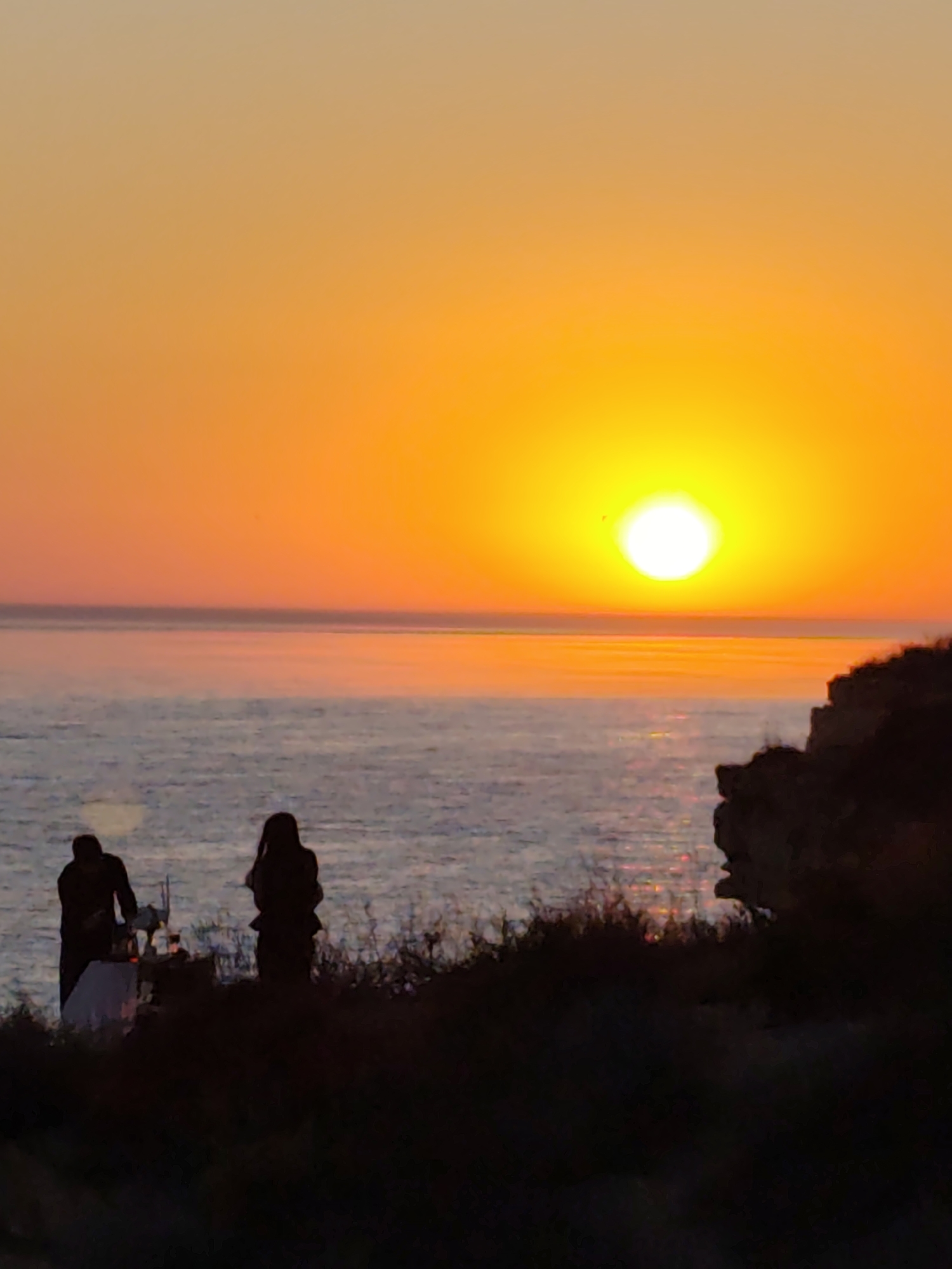 The image captures a serene sunset over the Caspian Sea in Aktau, Kazakhstan. The sky is ablaze with vibrant shades of orange and yellow, with the sun a bright, almost blinding disc near the horizon. The sea below reflects the warm hues of the sky, creating a shimmering expanse of light.
In the foreground, silhouetted against the dramatic sky, are two figures standing on a grassy cliff overlooking the water. To the left, a person appears to be tending to a small setup on the ground, possibly a picnic or a romantic arrangement with a white cloth and what looks like a bottle. The second figure stands slightly to the right and behind the first, gazing out at the sea. The figures are in silhouette, their features obscured by the backlighting, lending an air of quiet contemplation or shared intimacy to the scene.
The surrounding vegetation on the cliff edge is also in silhouette, creating a dark, textured foreground that contrasts with the bright sky and sea. A rocky outcrop is visible on the right side of the frame, adding a sense of ruggedness to the coastal landscape.
The time of day is clearly sunset, indicated by the position of the sun and the intense colors in the sky. The weather appears calm, with the sea being relatively still, allowing for the reflection of the sunset. The overall mood is tranquil and picturesque, evoking a sense of peace and natural beauty. There is no visible text in the image.
