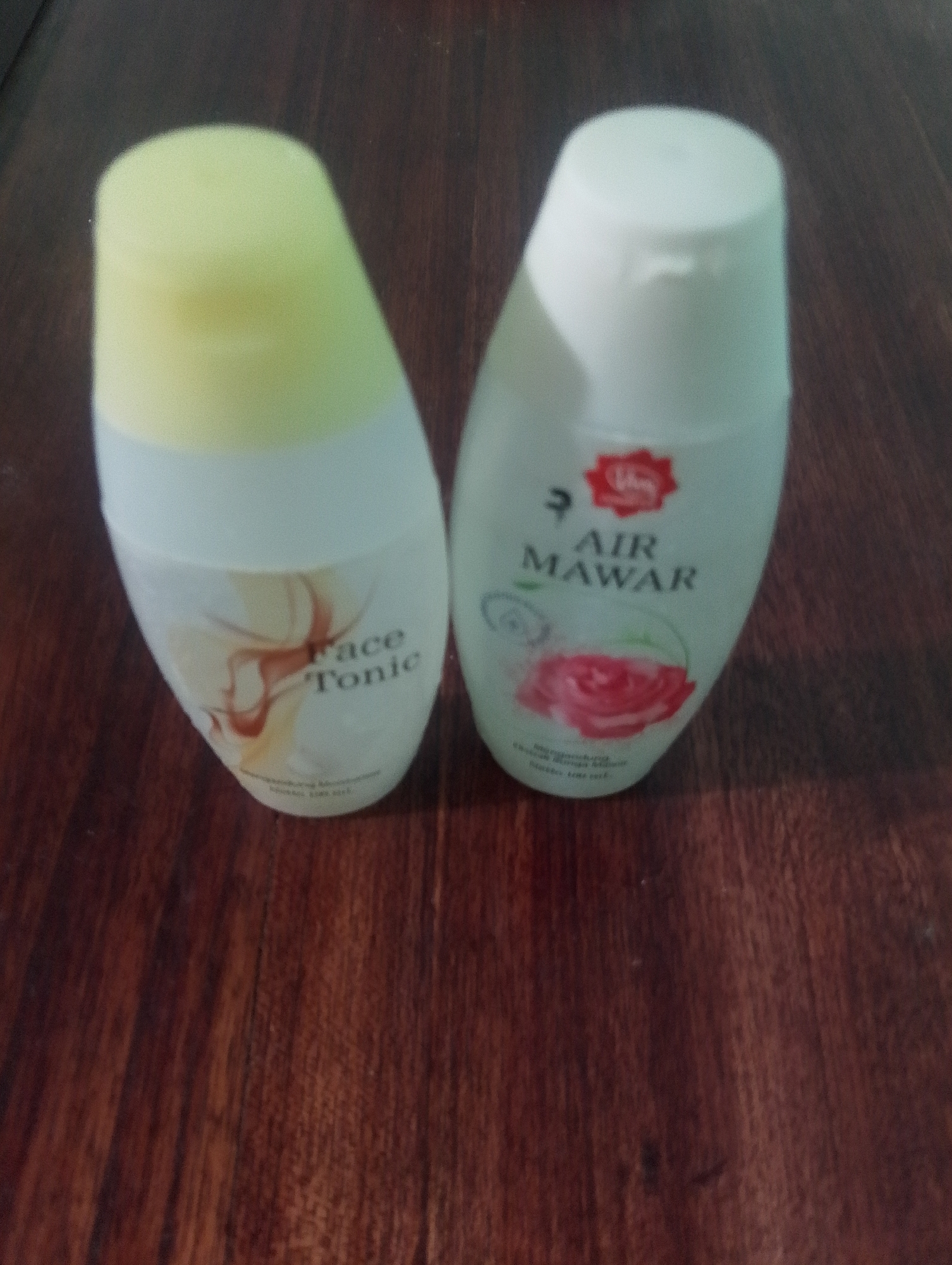 Two bottles of skincare products are displayed on a wooden surface. The bottle on the left is labeled "Face Tonic" and has a light yellow cap. The bottle on the right is white and labeled "Air Mawar" with a red rose graphic. The wooden surface appears to be a table or desk with a visible wood grain pattern. The lighting suggests an indoor setting, possibly a home. The location of Serbelawan, Indonesia is not evident from the image itself, but the products appear to be Indonesian brands.