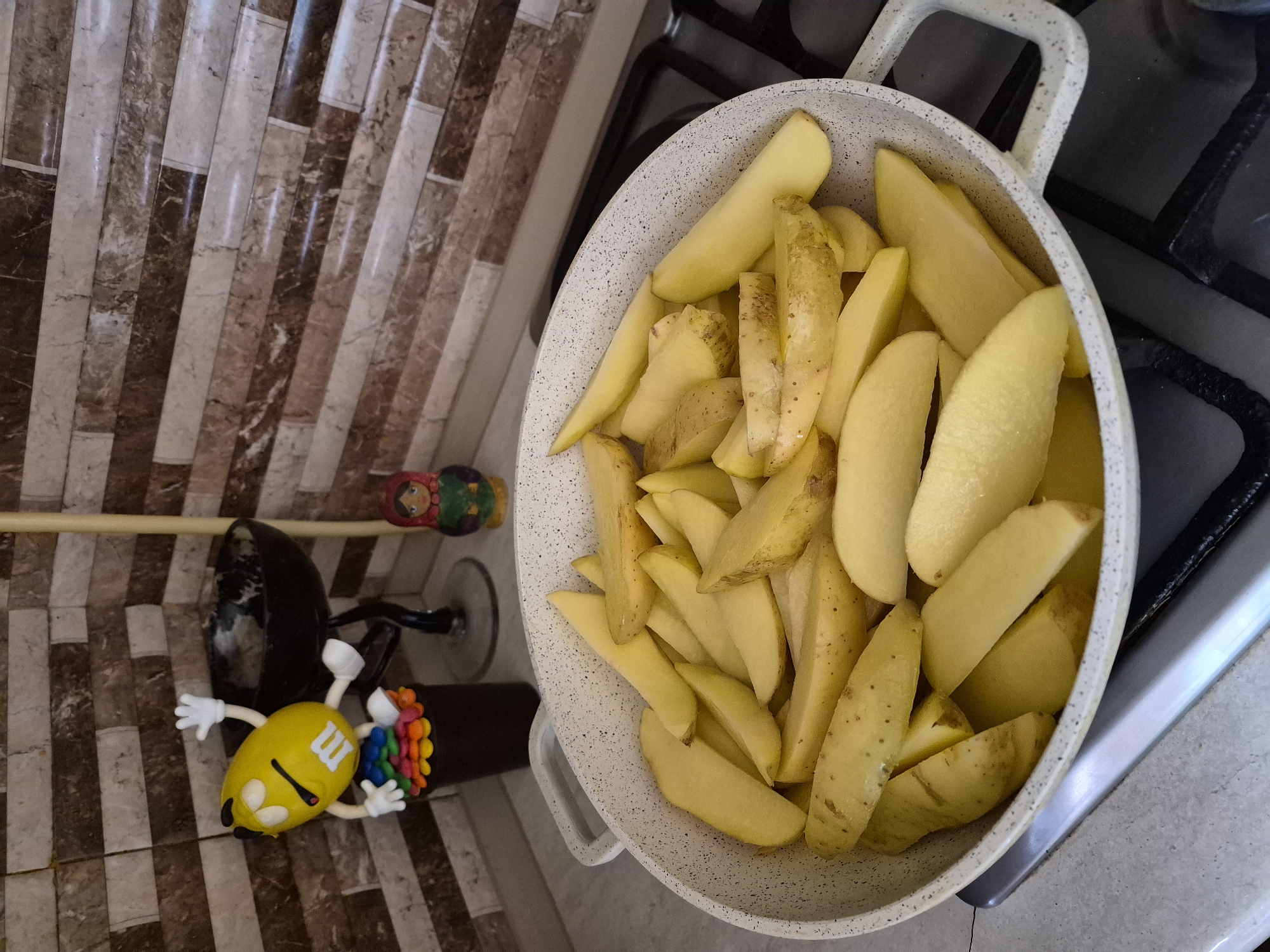 In a kitchen, likely in Yerevan, Armenia, a wide, shallow pan filled with cut potatoes sits on a stove. The potatoes are raw and have been cut into wedges. The pan is speckled white with gray handles. In the background, a patterned tile backsplash is visible. To the left of the pan, an M&M character figurine stands on a black pot, and further to the right, a small matryoshka doll is visible. The scene appears to be indoors, in a domestic setting.