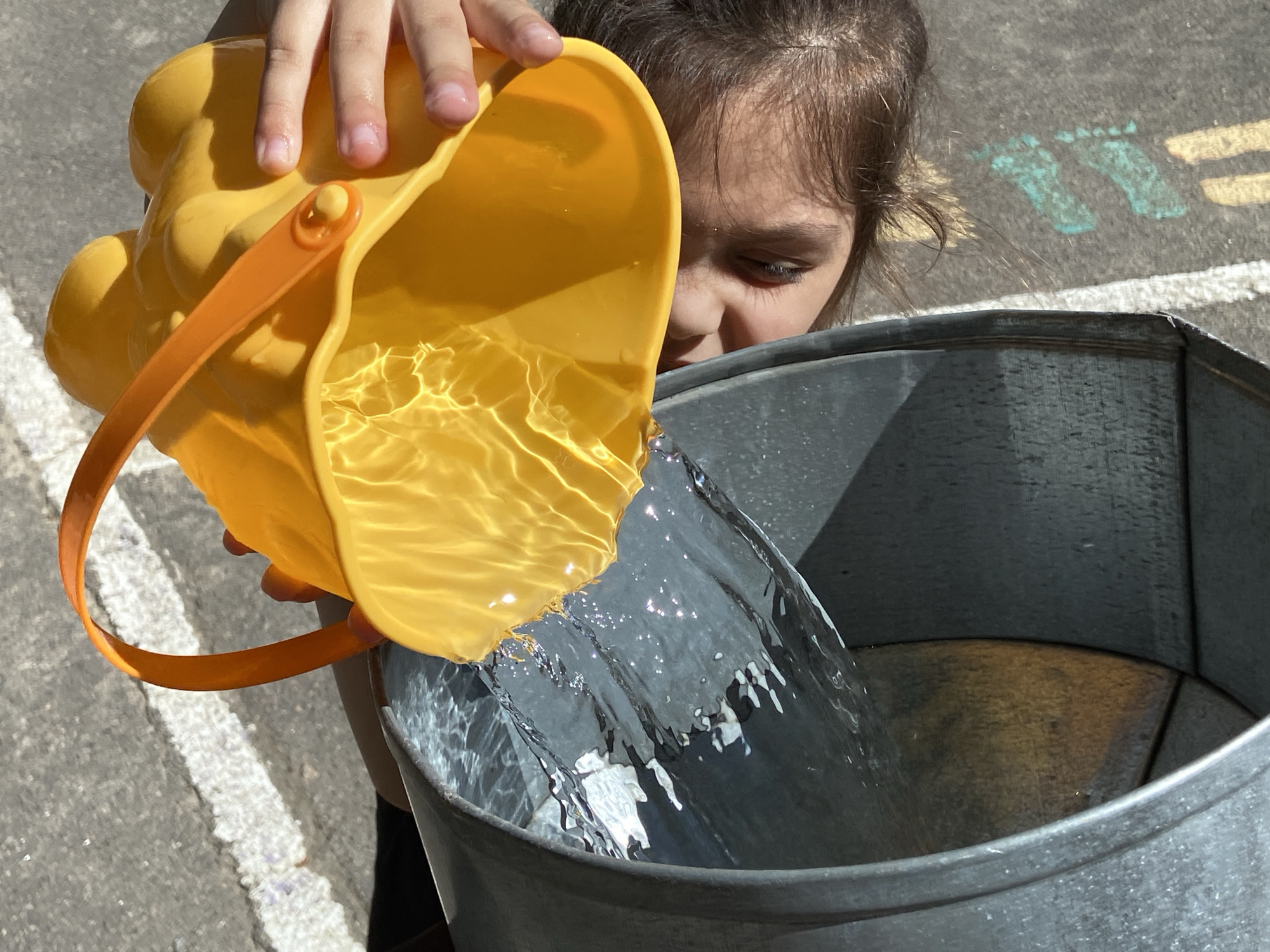 This is a close-up, outdoor shot of a young girl pouring water from a yellow toy bucket into a large, galvanized metal tub. The girl's face is partially visible, with her brown hair falling around her shoulders as she concentrates on the task. Her eyes are downcast, focused on the water stream. Sunlight illuminates the scene, casting shadows and highlighting the ripples on the surface of the water in the toy bucket and the spray as it enters the tub.
The background shows an asphalt surface with painted markings, including a pair of turquoise footprints and a yellow line. These markings suggest an outdoor play area, possibly a playground or a schoolyard. The time of day appears to be sunny, indicating fair weather. The overall mood is one of playful activity and concentration. The focus is on the action of pouring water, with the subjects and setting providing context for this simple, engaging moment.