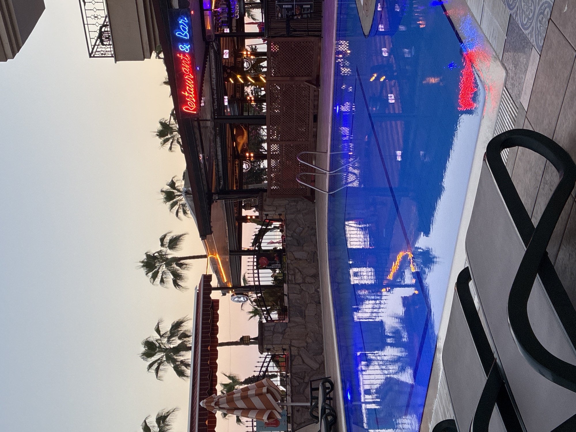 An outdoor evening scene depicts a hotel or resort swimming pool area in Alanya, Turkey. In the foreground, two dark-colored pool lounge chairs with black frames and fabric seats are partially visible. A rectangular swimming pool occupies the midground, filled with bright blue water reflecting artificial lights and the vibrant red and blue neon sign of a "Restaurant & Bar." Blue lights are illuminated within the pool, and a two-step metallic ladder extends into the water from the left edge. The pool deck is tiled in light tones, with some patterned sections.
Beyond the pool, a decorative stone wall topped with a black ornate metal fence separates the pool area from a restaurant/bar establishment. Behind this fence, a red and white striped umbrella is open, and a seating area with a red-striped awning is visible. The "Restaurant & Bar" features a prominent red neon sign spelling "Restaurant" and a blue neon sign for "& Bar." Inside, numerous warm string lights illuminate the space, and additional signs with text like "Where there's alcohol, there's conversation" and "We harvest good times together" are discernible. Wooden lattice panels are integrated into the lower structure of the restaurant. Bar stools and bottles are visible behind the bar counter.
The background features several palm trees silhouetted against a twilight sky, which transitions from a pale orange-pink near the horizon to a light blue overhead. A multi-story building with a dark-railed balcony is visible on the far right. No individuals are present in the image.