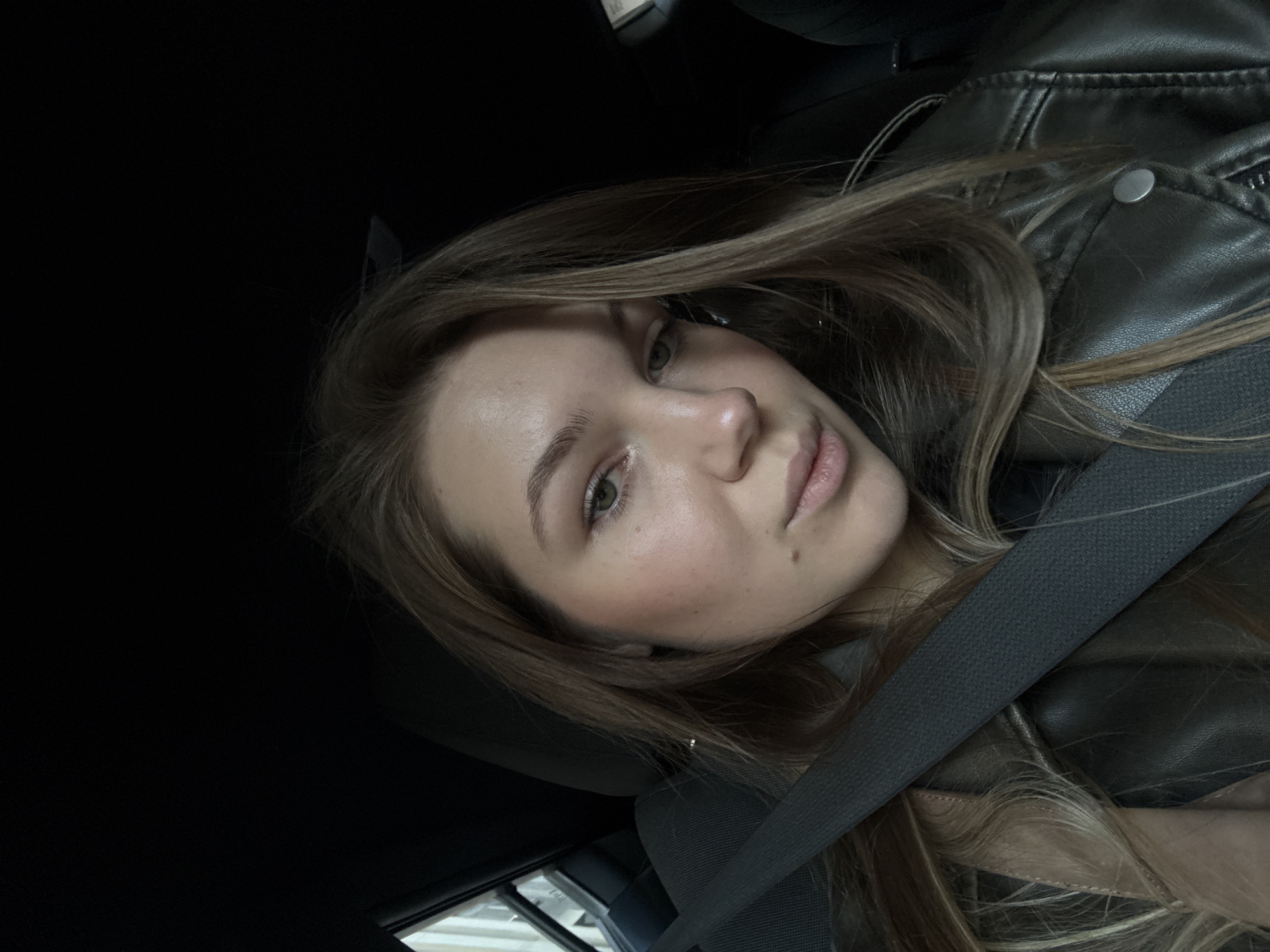 The image is a selfie of a young woman with long brown hair, wearing a black leather jacket and a seatbelt in a car. The setting appears to be inside a vehicle, possibly during daytime, as some light is visible through the window in the lower-left corner of the frame. The car is parked in an underground parking. The location is likely Belgrade, Serbia.