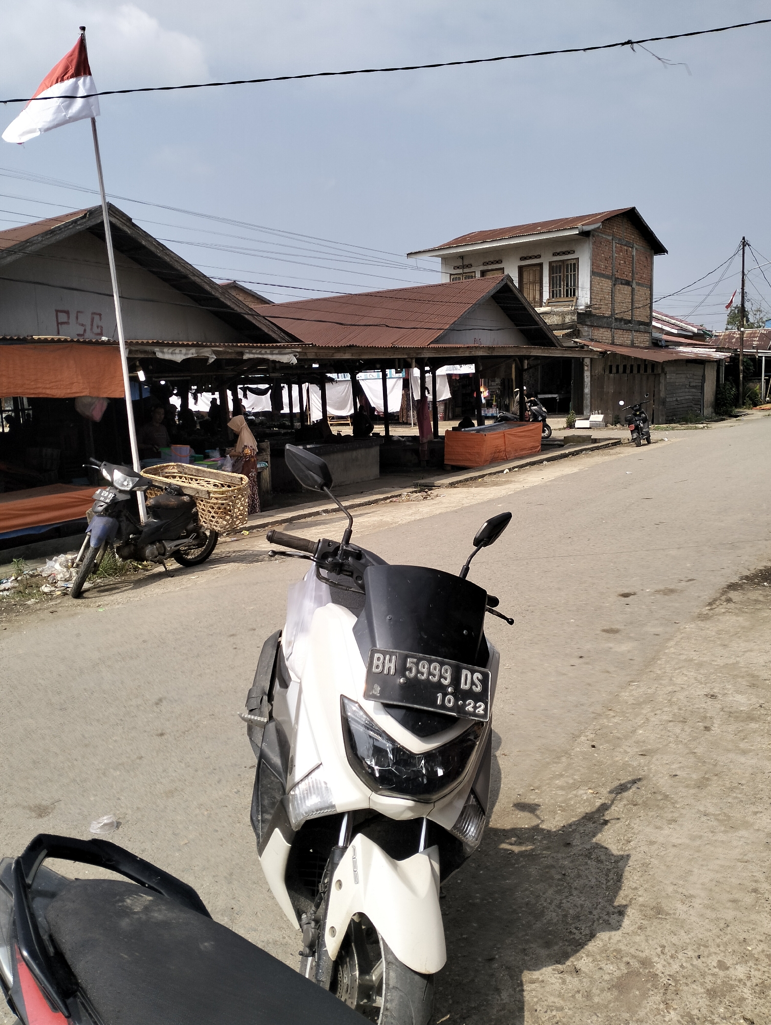 An outdoor street market scene is depicted in Serdang Jaya, Indonesia. In the foreground, the front of a white scooter is visible, with a license plate reading "BH 5999 DS 10-22". To the left of the scooter, the rear section of another scooter is partially visible. The background features a row of market stalls with tin roofs, some draped with orange tarpaulin. A flagpole with the Indonesian flag (red and white) flies prominently on the left side of the frame. Several people are present within the market stalls, some appearing to be vendors. A dark-colored scooter with a large woven basket is parked near the stalls. Beyond the market, a two-story building with brick and white plaster construction stands, and power lines crisscross the sky. The ground is paved with asphalt, and sunlight casts shadows.