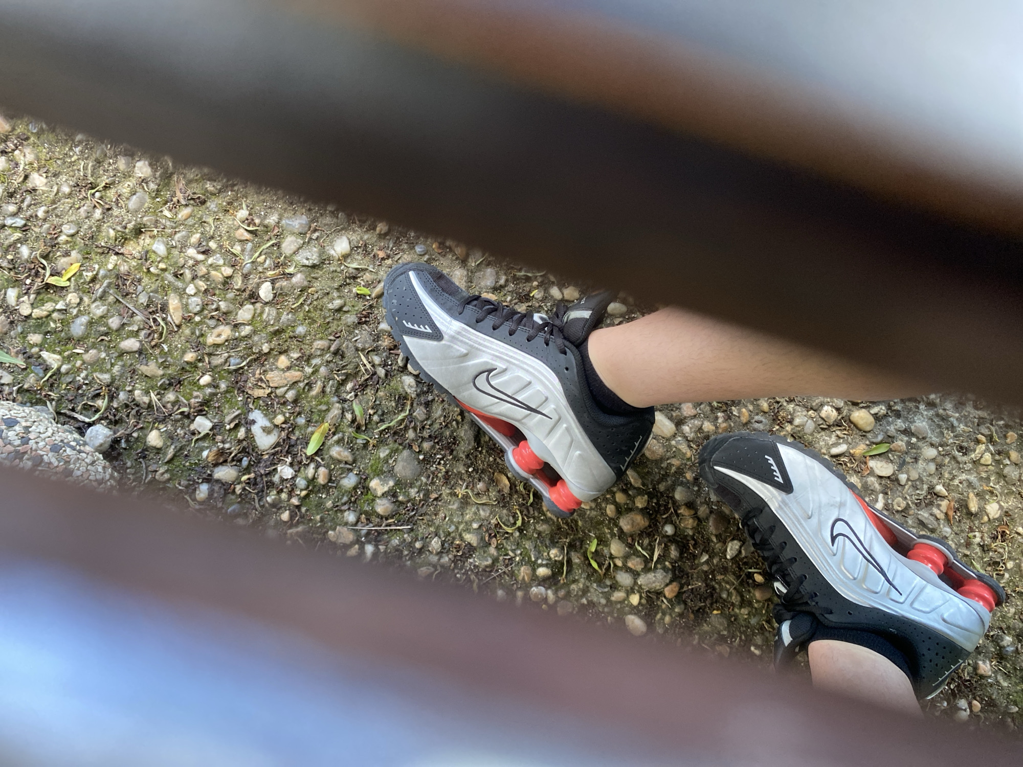 Two feet wearing black and silver Nike Shox sneakers are visible on a gravelly ground. The sneakers have red cushioning visible in the heel. The ground is a mixture of small grey and brown stones, with patches of green moss and some dried leaves and twigs scattered throughout. A dark, blurry horizontal shape, possibly a railing or bench, partially obscures the upper portion of the image. The lighting suggests it is daytime, and the presence of moss and sparse vegetation might indicate an outdoor location. The angle of the shot, looking down at the feet and the ground, gives a first-person perspective. There is no visible text in the image.