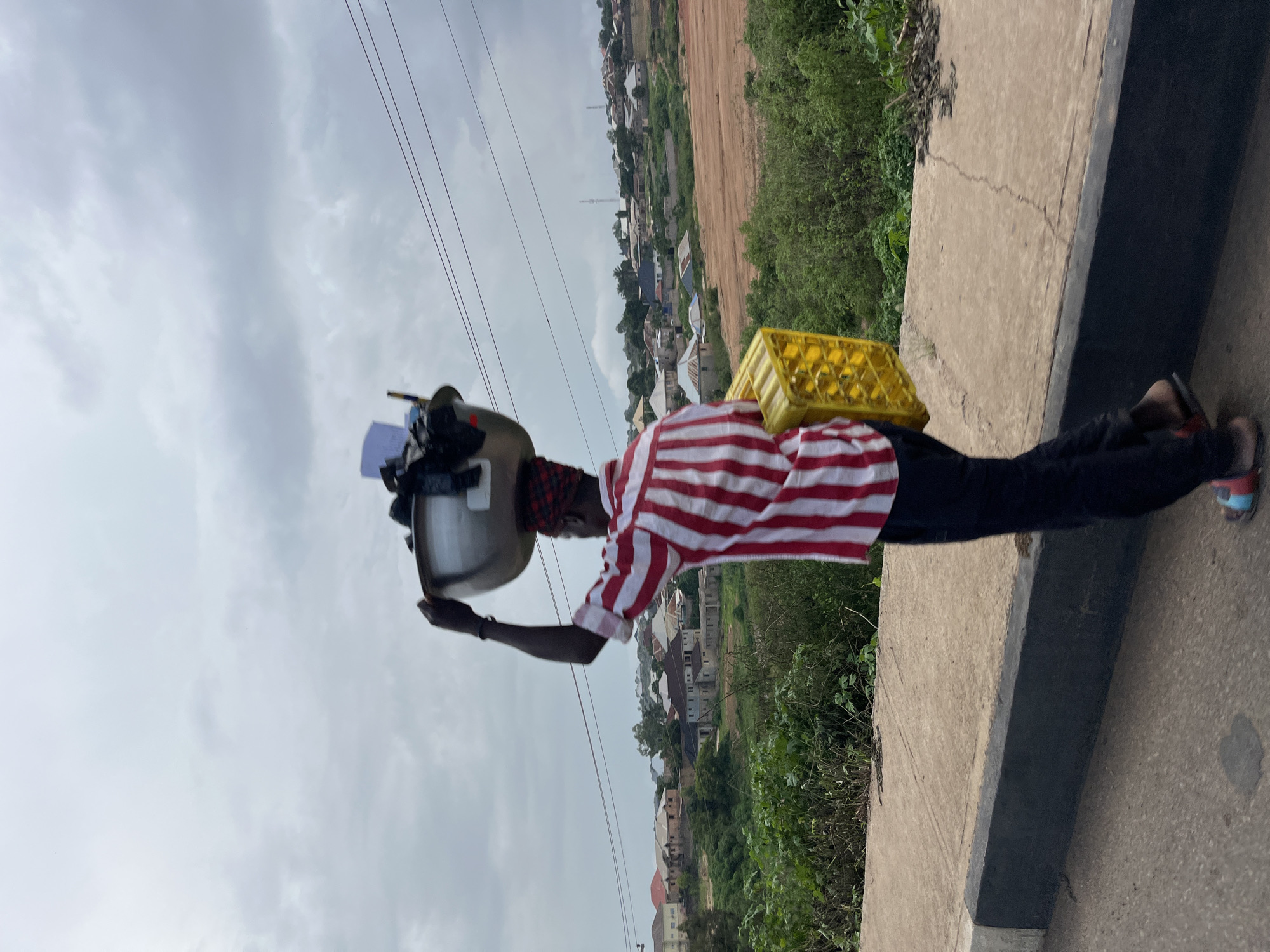 A person walks along a paved road, balancing a large metal basin filled with various items on their head, while carrying a yellow plastic crate in their right hand. The individual is wearing a red and white striped shirt and dark pants. The background reveals a residential area with buildings, lush green vegetation, and a cloudy sky. The location is identified as Gura Top, Nigeria.