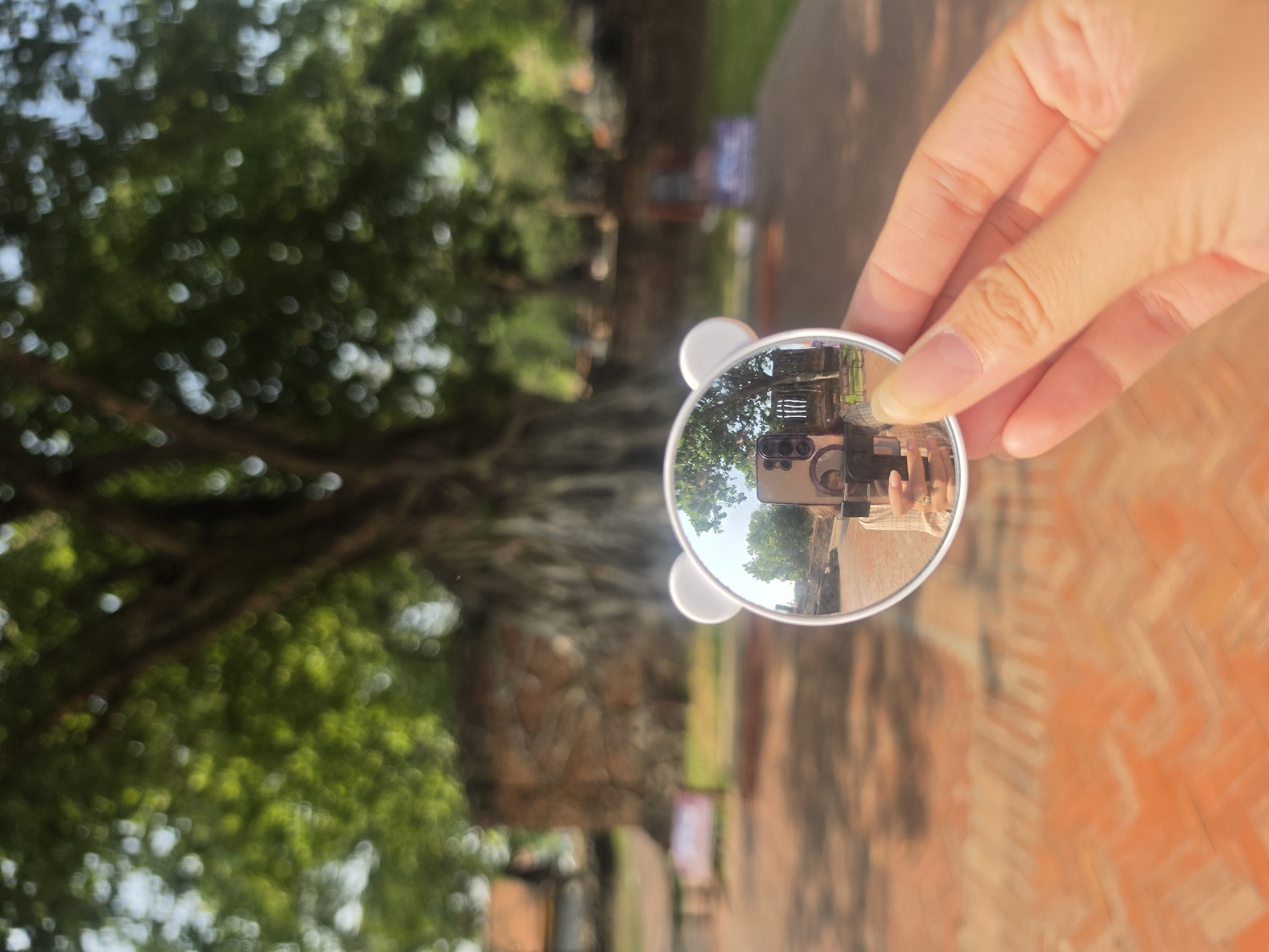 A close-up, rotated 90 degrees clockwise, shows a fair-skinned hand holding a small, circular mirror with a white frame, possibly adorned with small, rounded protrusions resembling cat ears at the top. The mirror prominently reflects a modern smartphone camera with multiple lenses, and partially, the face of the photographer, indicating a creative self-portrait or a reflection shot. Within the mirror's reflection, an outdoor scene is visible, featuring green foliage, a clear sky, and what appears to be an old brick wall or building.
The background of the main image is intentionally blurred, creating a bokeh effect. It consists primarily of lush green tree canopy and branches on the left, suggesting large, mature trees, possibly with visible trunks or roots. Towards the right and bottom of the frame, blurred orange-brown brickwork is visible, hinting at an ancient pathway or building foundation. The lighting is bright and natural, indicating a clear, sunny daytime. The scene evokes an atmosphere of exploration at a historical or natural outdoor site, consistent with Phra Nakhon Si Ayutthaya City Municipality, Thailand. There is no visible text in the image.