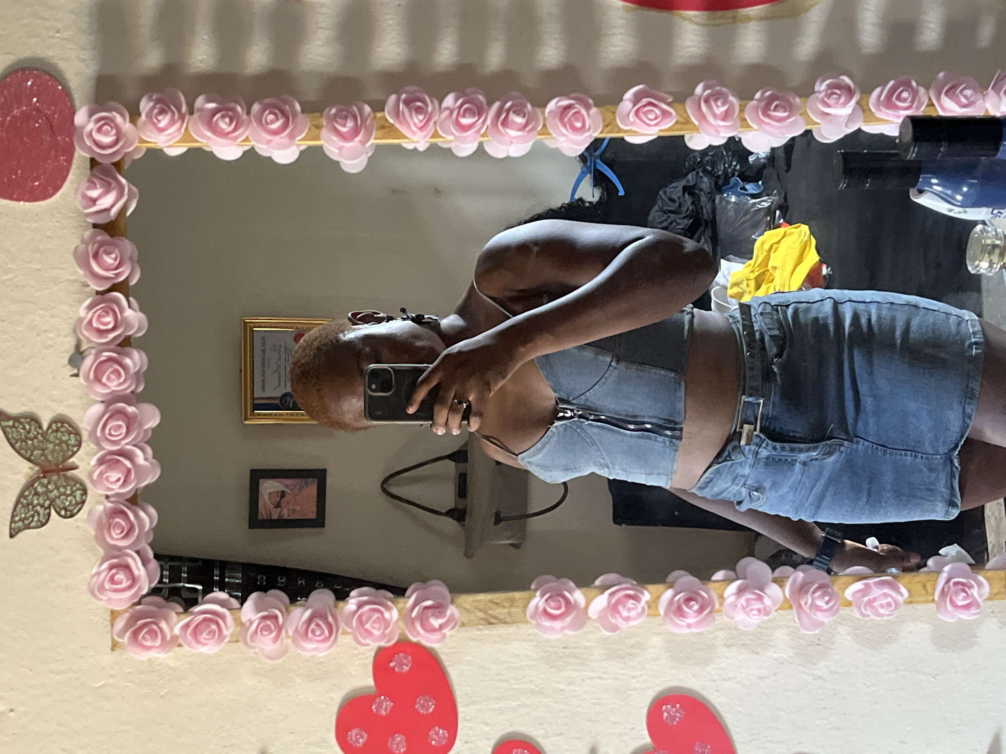 The image, rotated 90 degrees counter-clockwise, captures a person taking a mirror selfie indoors. The individual, appearing to be female, has dark skin and short, reddish-brown hair. They are wearing a light denim-colored sleeveless crop top or bustier with a front zipper, paired with matching denim shorts or a skirt. A black smartphone with a triple camera setup is held up to capture the reflection, obscuring most of the person's face. A silver ring is visible on the left ring finger, and a dark-colored watch is on the left wrist.
The mirror features a decorative frame adorned with numerous light pink, rose-shaped embellishments. On the wall to the left of the mirror, a red, glittery oval decoration and a gold-toned butterfly ornament are visible. Below the mirror, several red, glitter-speckled heart decorations are affixed to the wall.
Reflected in the mirror, the background reveals an indoor setting with light-colored walls. A golden-framed object, possibly a certificate or picture, is partially visible on the wall, along with a smaller, dark-framed picture featuring a figure. On a surface behind the person, various items are reflected, including black plastic bags, a bright yellow cloth, and bottles (one dark blue, one clear with a gold top). Wires and what appears to be a power outlet or strip are also visible on the wall. A black object, possibly a remote control, with faint numbers like "88" visible, lies on a surface.
The activity is the act of taking a mirror selfie in what appears to be a personal space, possibly in Jalingo, Nigeria. The time of day is not explicitly clear but is likely either daytime or evening with artificial indoor lighting.