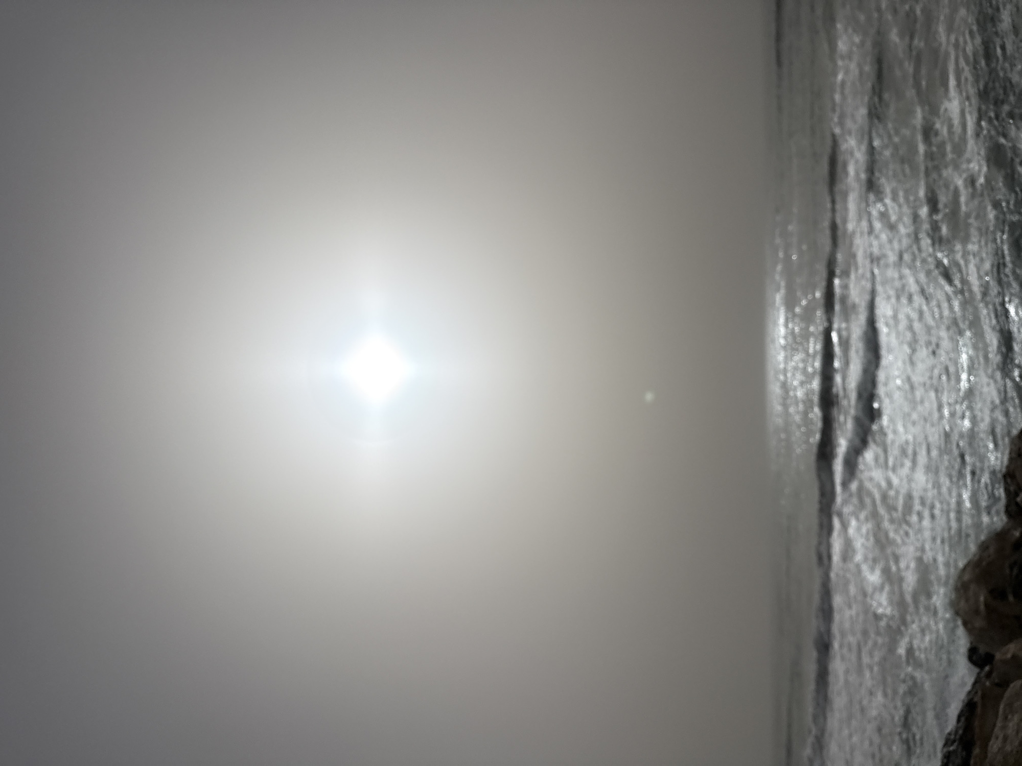 The image depicts a shoreline obscured by fog. A bright light source, presumably the sun, is visible through the mist. Water with rough currents is visible on the right side of the image, with rocky outcroppings in the lower right corner. This is likely a seaside scene in Tel Aviv, Israel.