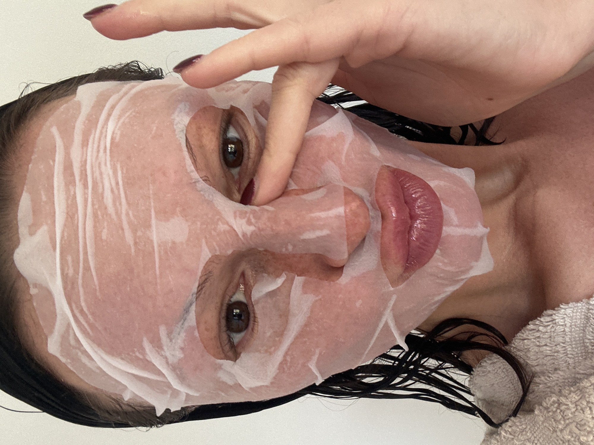 A female subject is wearing a translucent facial mask, adjusting it with the index finger of her right hand. Her dark, wet hair is visible around the mask and at her neck. Her eyes, visible through the mask openings, are brown. Her lips are exposed and appear glossy. The background is a light, neutral color. The subject is in a slightly rotated, close-up shot. There is no evidence to suggest this image was taken in Tunbridge Wells, United Kingdom.