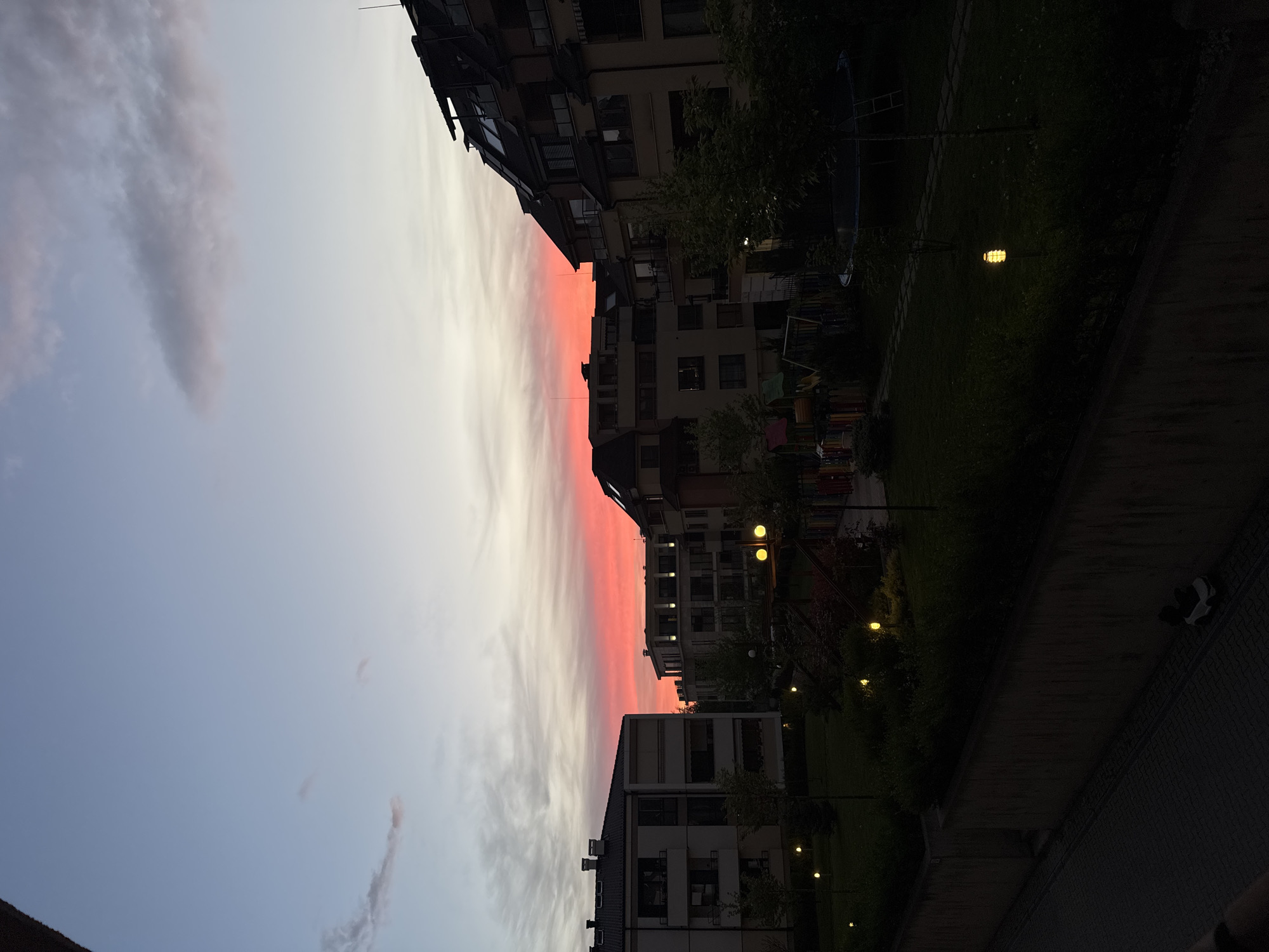 Captured at dusk, the scene showcases residential buildings in Sofia, Bulgaria. The sky exhibits a gradient of light blue to a reddish-pink hue, likely indicative of a sunset or sunrise. Buildings of multiple stories line the right side of the frame, with visible windows and varying architectural styles. Street lights illuminate the surroundings, indicating that it is nighttime. A park area with vegetation and playground equipment can be seen between the buildings. The foreground shows a dimly lit sidewalk with some dark objects that look like people sitting on a bench, further emphasizing the evening setting.
