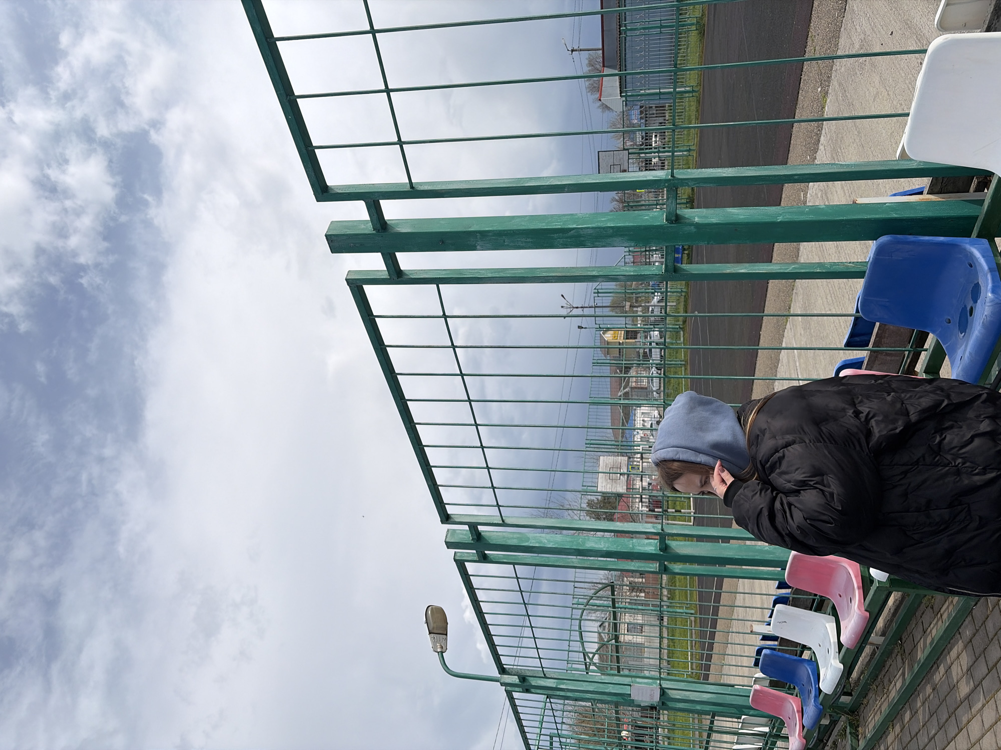 A young woman wearing a light blue hooded sweatshirt and a black jacket is sitting on a row of colorful stadium seats behind a green fence. The seats are arranged in a sequence of pink, blue, white, and pink. Beyond the fence, there is a street and several buildings. A street lamp is attached to the fence. The sky above is cloudy and overcast. The location is Novopokrovskaya, Russia.