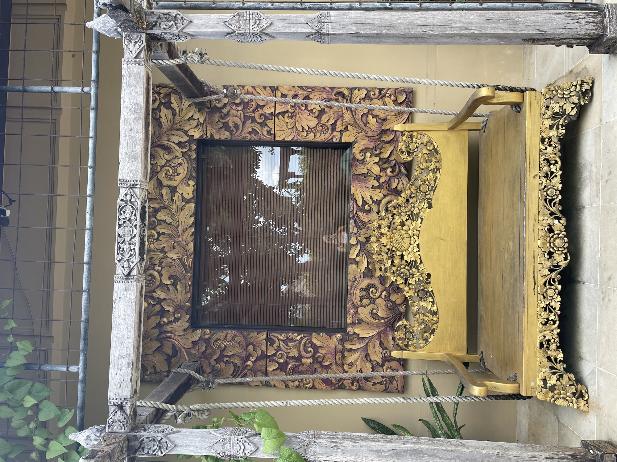 The image shows an outdoor area in an unknown city. A wooden frame supports a swing with intricately carved floral designs in gold and purple. A window with blinds is visible behind the swing, and plants are growing nearby. The area appears to be a courtyard or garden setting.
