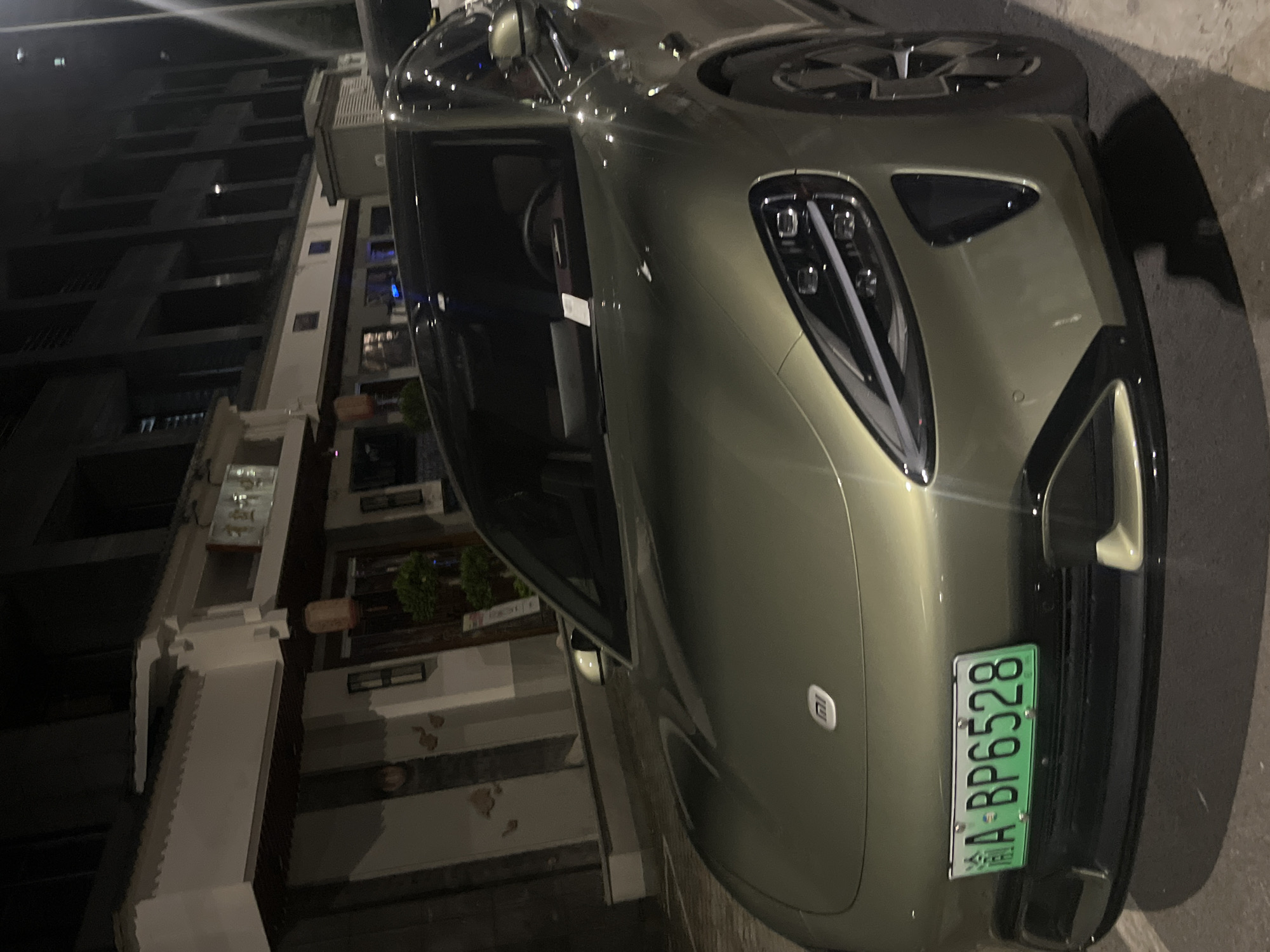 A green license plate reading "A-BP6528" is affixed to the front of a metallic green car parked on a street at night in Jiulongpo District, China. The car's hood features a prominent silver emblem. In the background, a building with traditional Chinese architectural elements, including a white facade, dark wood trim, and red lanterns, is visible. Adjacent to this building is a modern, dark building with a grid-like facade. Streetlights illuminate the scene, casting shadows on the pavement.