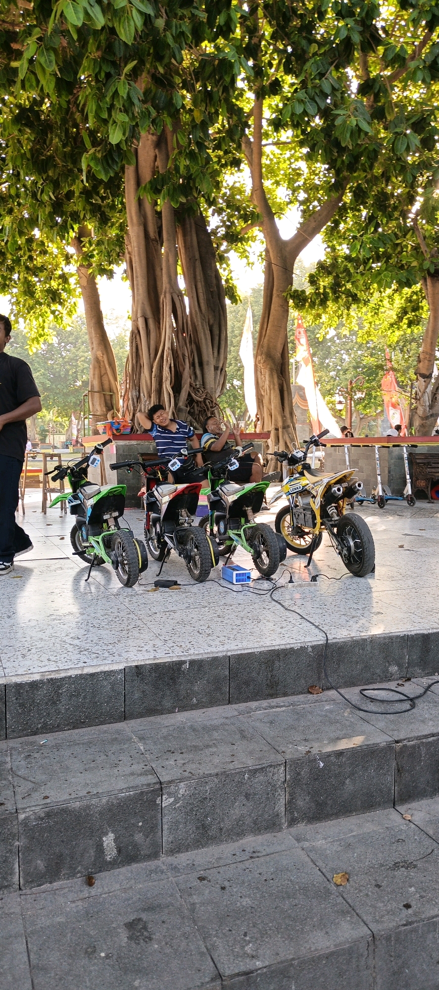 In a public park in Klaten, Indonesia, beneath the shade of a large banyan tree, several children's dirt bikes are parked in a row on a tiled surface. The bikes are various colors, including green, red, and yellow, and appear to be electric. A blue battery charger and power strip are visible, connected to one of the bikes, suggesting they are being charged. Two men are seated on a bench behind the bikes. One man is wearing a blue and white striped shirt and appears to be relaxing, with his head leaning back. The other man, in a yellow and black striped shirt, is holding up two fingers as if gesturing or posing for a photo. A man in a black shirt and dark pants stands to the left of the frame, partially obscuring one of the green bikes. In the background, flags are displayed, and the sunlight filters through the leaves of the banyan tree, creating a dappled light effect. The scene suggests a casual outdoor gathering or a place where children can rent and ride these small bikes.