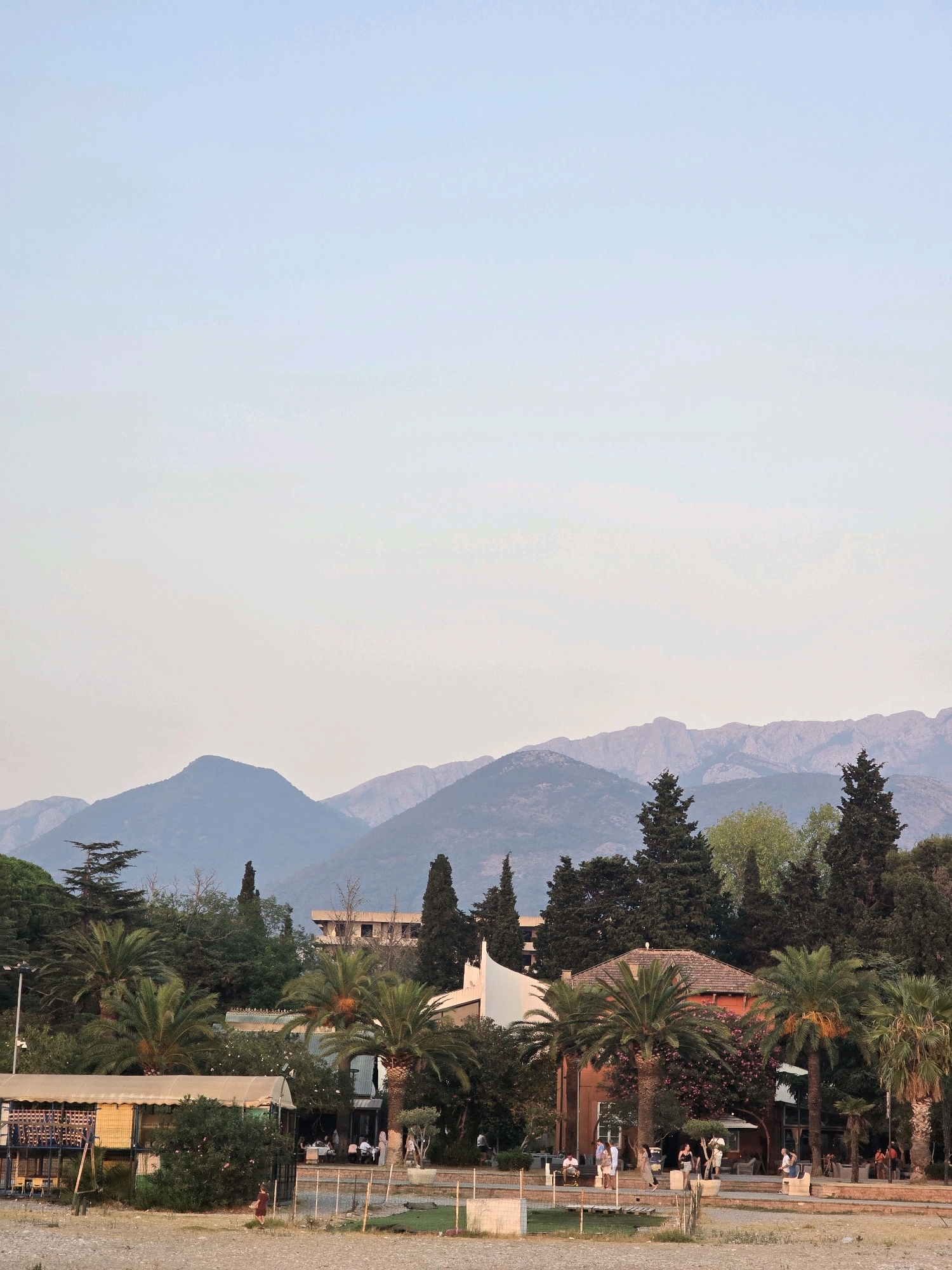 This image captures a coastal scene with a backdrop of majestic mountains, likely the Rumija Mountains, near the city of Bar, Montenegro. In the foreground, a sandy beach transitions into a paved walkway lined with palm trees and sparse vegetation. A building with a red-tiled roof, featuring several large windows and a covered patio, is visible, with people seated at tables outside. Adjacent to it, a colorful structure, possibly a cafe or kiosk, is situated. Further back, several taller buildings and numerous trees, including cypress and palm varieties, dot the landscape, creating a lush, green environment. The overall atmosphere suggests a relaxed, late afternoon or early evening setting, with the soft light casting a warm glow over the scene. The sky is a pale, hazy blue, indicating fair weather.