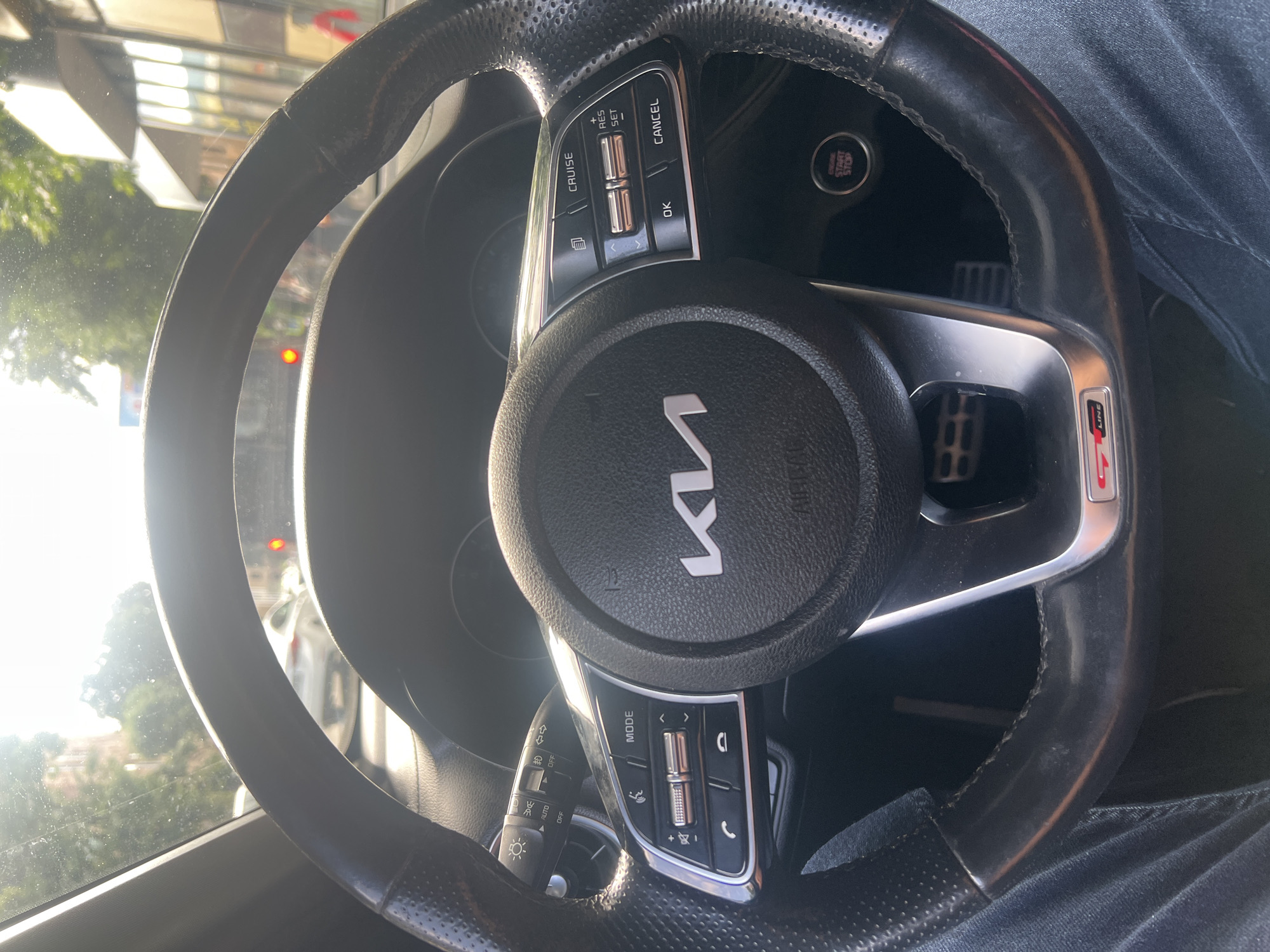 The image shows the interior of a car, focusing on the steering wheel and dashboard. The steering wheel is black with textured grips and features several control buttons on its left and right sides. On the left, there are buttons labeled "CRUISE", "RES", "+", and "SET", along with two silver metallic toggles. On the right, buttons for "MODE", "<", ">", a phone icon, and another phone icon are visible, along with a silver metallic roller. A prominent "KIA" logo is centered on the steering wheel. To the right of the steering wheel, a circular "ENGINE START STOP" button is visible. A red "GT LINE" badge is present on the lower right side of the steering wheel's spoke. The scene appears to be from the driver's perspective, with part of the driver's leg, clad in denim jeans, visible in the lower right corner. The background outside the car, visible through the windshield, shows sunlight filtering through trees and hints of buildings, suggesting an outdoor setting during daytime. The overall lighting indicates it might be a sunny day. The location context provided is Vanadzor, Armenia. The image captures a detail of the car's interior and its controls.
--- OCR Start ---
AUTO
OFF
OFF
MODE
>
>
LINE
旬
CRUISE
>
OK
START
+
RES
SET
CANCEL
--- OCR End ---