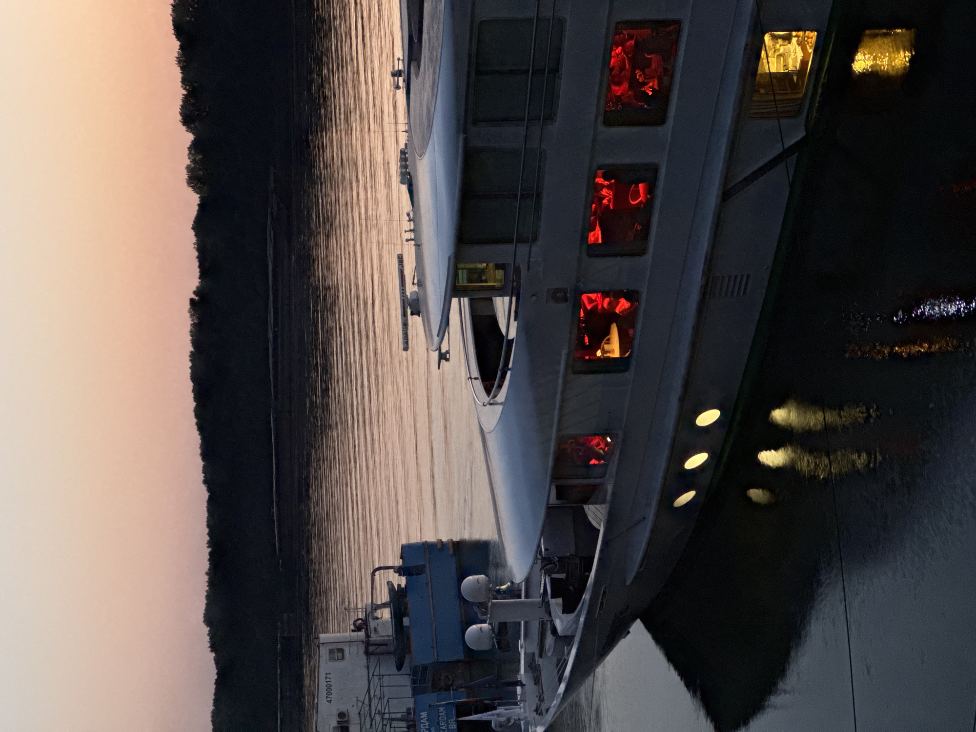 The image depicts a large, multi-deck boat docked on the Danube River at dusk in Ruse, Bulgaria. The sky is a gradient of soft pinks and oranges. The boat's windows glow with warm, red light, suggesting activity inside, possibly a restaurant or lounge. Several smaller circular windows along the lower hull also emit a yellow light, casting reflections on the dark water. In the background, another smaller blue boat is visible, along with a more distant, low-lying forested shoreline. The water appears calm, with subtle ripples and reflections of the boat's lights. The overall atmosphere is peaceful and serene, capturing the transition from day to night.