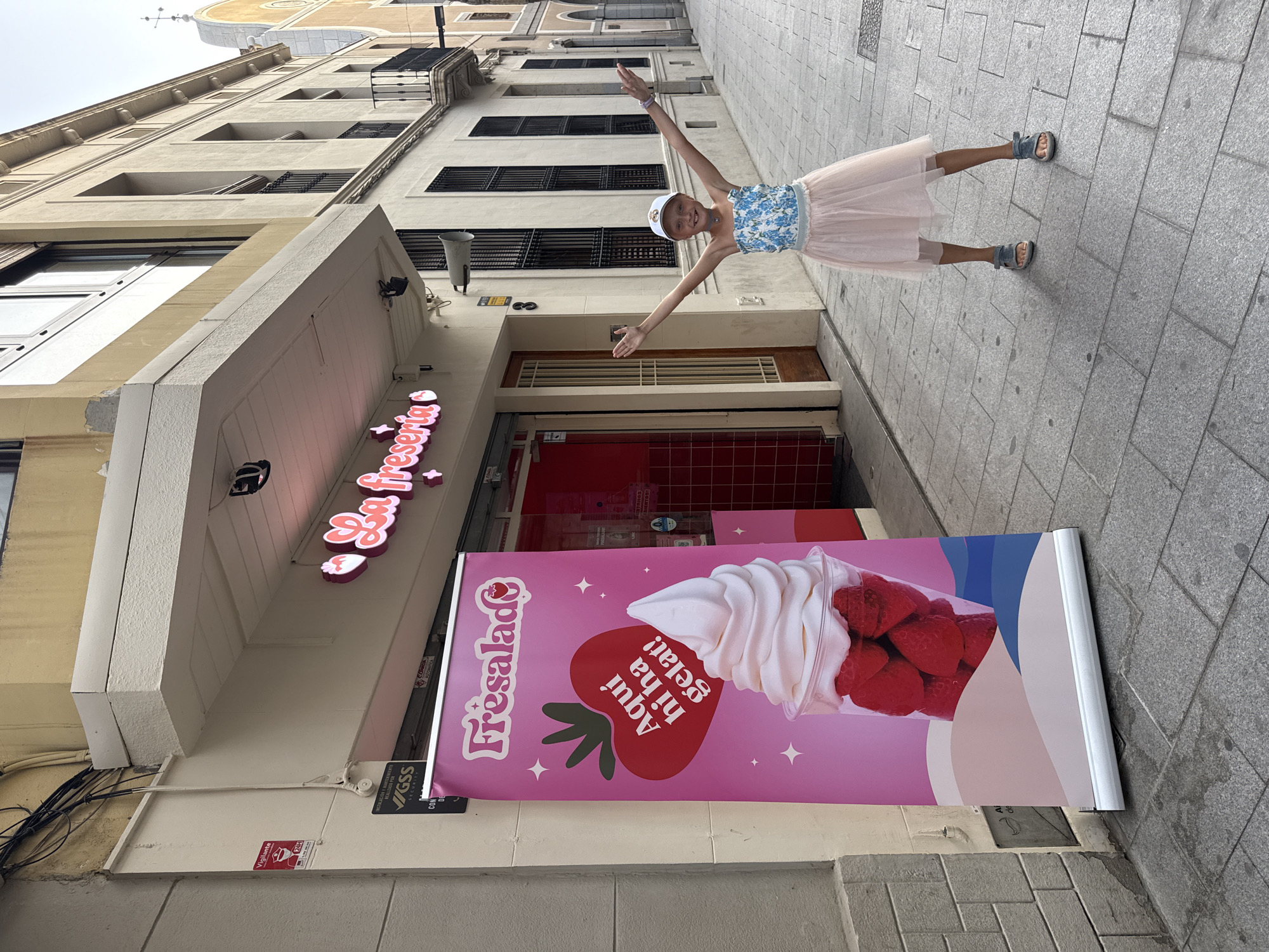 A young girl, wearing a floral blue and white strapless top, a light pink tutu skirt, a white baseball cap, and gray sandals, stands on a paved sidewalk with her arms outstretched in front of a store with a pink awning. The store, identified by a neon sign that reads "La Fresería" and a large pink banner displaying an image of a strawberry ice cream cone, is located in Mataró, Spain. The banner features the text "Aquí hi ha gelat!". The girl is the sole subject of the image. The environment is urban, with buildings lining the street.
