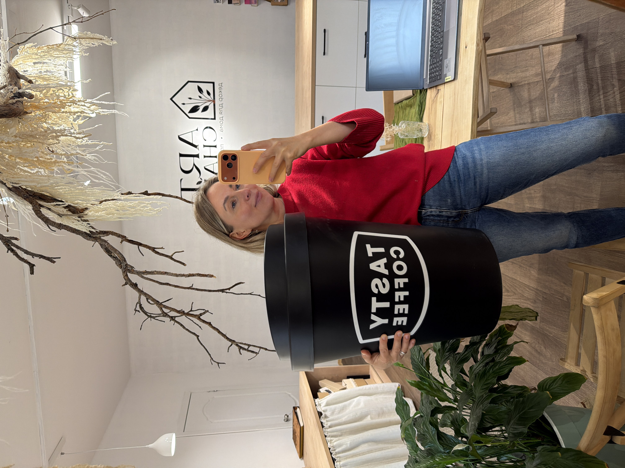 A woman stands in a room in Chaikovsky, Russia, holding a large black container labeled "TASTY COFFEE." She wears a red sweater and jeans and is taking a selfie with an orange phone. The room features a desk with a laptop, a faux plant, and decorative branches. There is a sign on the wall with the text "ART CHA" in Russian. The room has white walls and bright lighting.