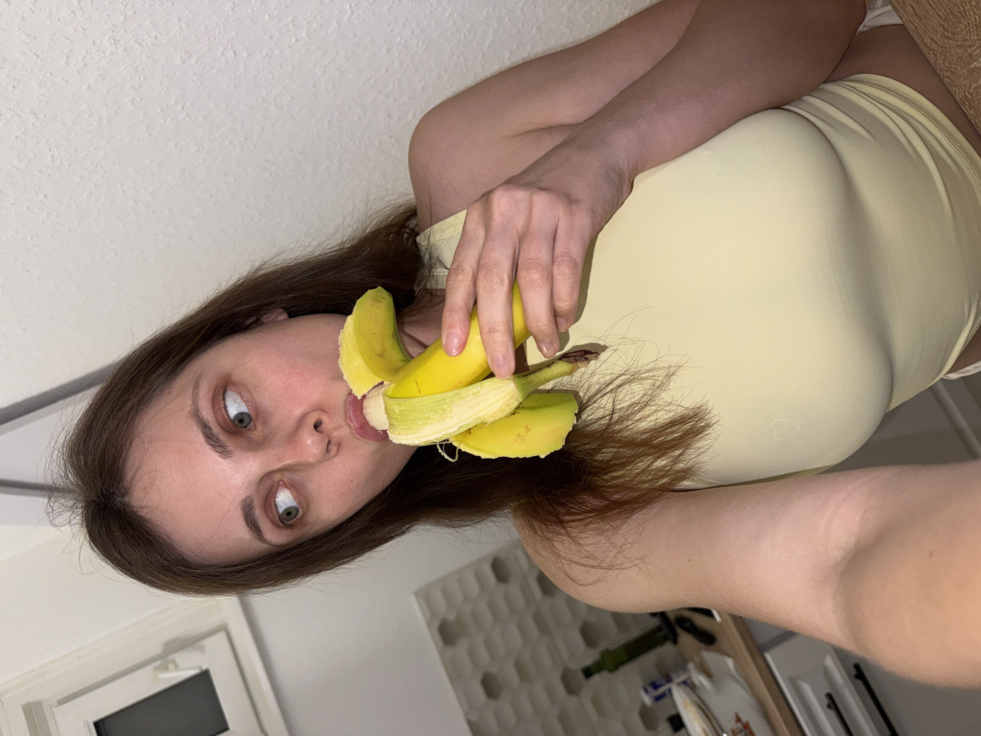 A woman is holding a peeled banana to her mouth. She has brown hair and fair skin, and is wearing a yellow top. The background includes a wall and a glimpse of a kitchen counter. The location is not discernable from the image and cannot be identified as Kőröshegy, Hungary.