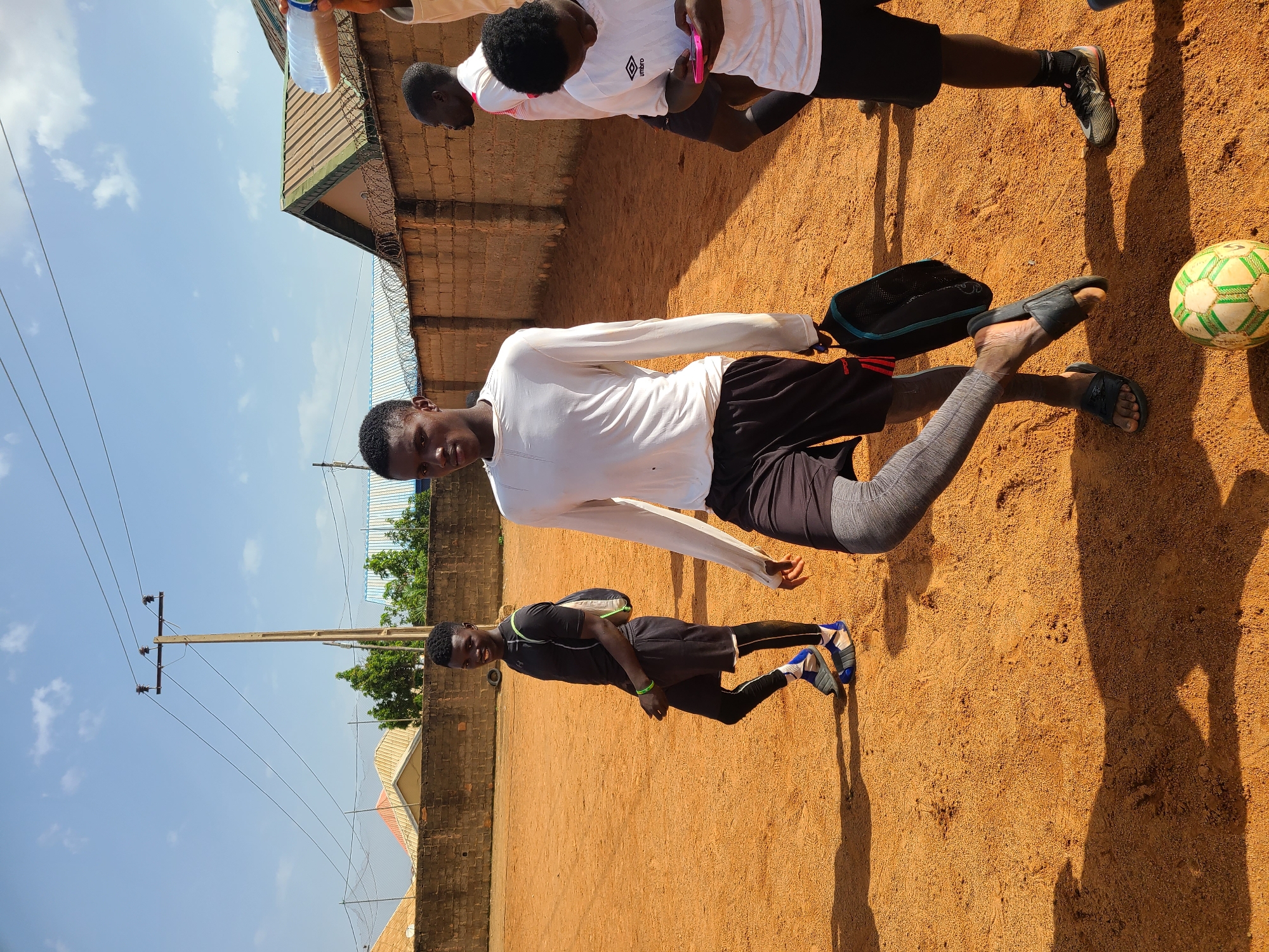 The image depicts a group of young men on a dirt field in Barakin Lerawi, Nigeria. The sun is shining brightly, casting distinct shadows on the ground, suggesting it is midday. The sky is a clear blue with a few scattered white clouds.
In the foreground, a young man wearing a white long-sleeved shirt and black shorts is posing, looking directly at the camera. He is positioned in the center of the frame, with his left leg slightly bent. To his right, another young man in a black t-shirt, black shorts, and leggings is standing, also looking towards the camera. He is carrying a backpack over his shoulder. Further back, partially visible, are two more individuals. One is wearing a white t-shirt with the word "Umbro" visible on it, and black shorts. The other individual is only seen from the chest up, wearing a white t-shirt.
On the ground near the center-right of the image, a soccer ball with green and white hexagonal patterns is visible. Adjacent to the soccer ball are two black bags. The background consists of a brick wall and a corrugated metal roof structure, indicative of a building. In the distance, a utility pole with electrical wires stretches across the frame, and lush green trees are visible. The overall atmosphere appears casual and sporty, suggesting a break or a moment captured during a game or practice.