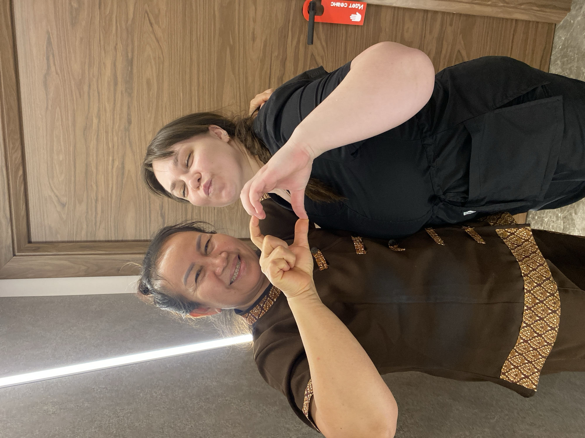 Two women are posing together indoors, likely at a spa or massage salon. The image is oriented vertically, showing the subjects standing side-by-side. On the left, a woman with fair skin and dark hair, tied back, wears a black top. She makes half of a heart shape with her hands and playfully puckers her lips. Beside her, a woman with East Asian features and dark hair, tied in a bun, is dressed in a dark brown uniform with distinctive gold patterned trim, possibly traditional Thai attire. She completes the heart shape with her hands and smiles warmly at the camera.
The background features a wooden door on the right, which has a red door hanger with a white hand icon and the Russian text "Идет сеанс," meaning "Session in progress" or "Massage in progress." This text, combined with the traditional attire of one of the women, strongly suggests the setting is a Thai massage or spa in Votkinsk, Russia. To the left, a grey wall features a horizontal strip of light. The overall mood is friendly and joyful, capturing a moment of connection between the two individuals.