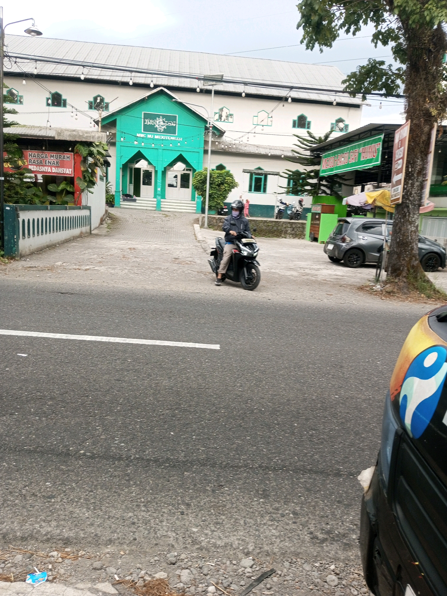 The image depicts a street scene in Garung, Indonesia, in front of a mosque. A man on a black motorcycle is in motion, moving away from the camera. He is wearing a dark jacket, light-colored pants, and a helmet. To the right of the road, there is a grey car parked. Further to the right, a large tree with a textured bark stands. In the background, a white building with green accents, identified by signage as "Masjid NU Mojo Tengahl," is prominent. A red sign with white text, reading "HARGA MURAH RASA ENAK PEDASNYA DAH SYAT," is visible on the left side of the image. The road is paved, with a white line marking the lane. The environment appears to be a mix of urban and natural elements, with buildings, a road, vehicles, and vegetation.