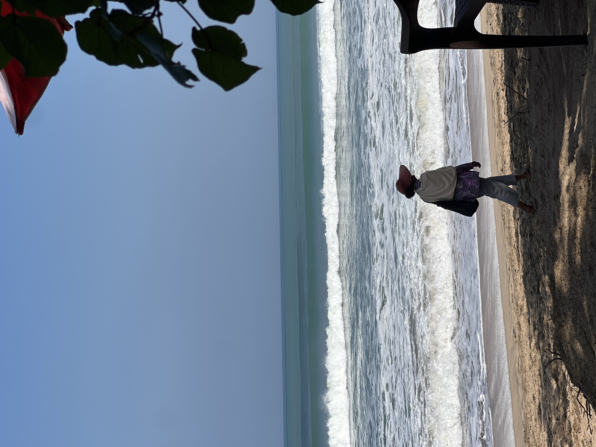 A lone figure walks along the beach in Kuta, Indonesia. The person, facing away from the camera, wears a wide-brimmed red hat, a light-colored shawl draped over their shoulders, and light-colored pants. They carry a dark bag over their left shoulder. The individual is walking on the wet sand near the water's edge, with gentle waves breaking on the shore. The ocean stretches out to the horizon under a clear blue sky. In the foreground, the sand is dappled with shadows from unseen foliage, and a dark plastic chair is partially visible on the right.