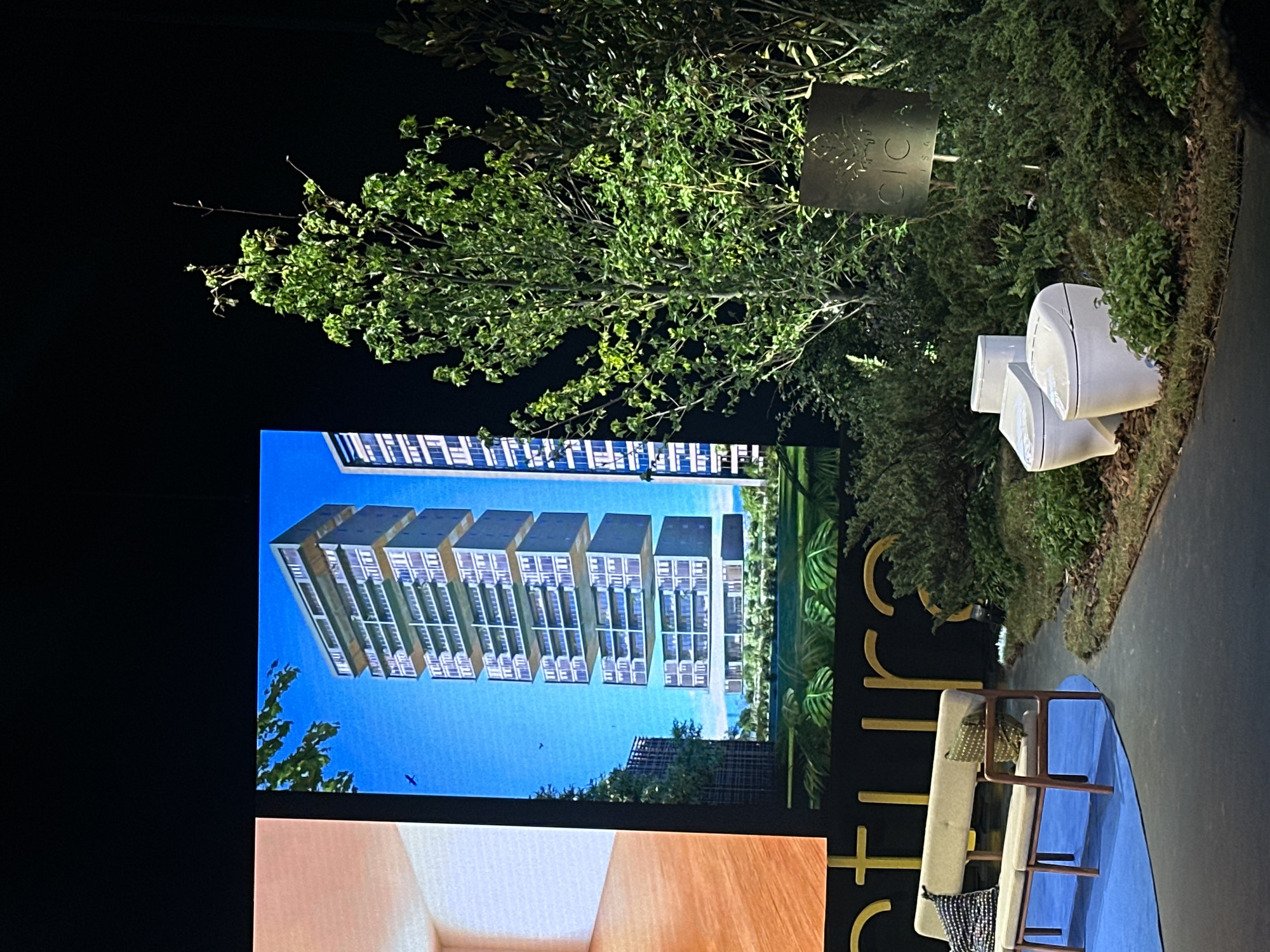 The image captures an outdoor or semi-outdoor display area at night, likely for a real estate development or architectural project in Huixquilucan de Degollado, Mexico. No people are visible.
The scene is dominated by a large, upright digital screen on the left, showcasing architectural renderings. The upper part of the screen displays a modern, multi-story building characterized by stacked, rectangular volumes, each featuring numerous balconies or outdoor terraces, set against a bright blue sky. A small bird can be seen in the sky. The lower part of the screen shows a close-up of what appears to be an interior space, with warm wooden tones.
To the right of the screen, dense, lush green trees and bushes are illuminated, suggesting artificial lighting against the dark night sky. A square, dark metal sign with a cut-out stylized leaf or tree logo is partially obscured by the foliage. On the sign, the word "CIO" is clearly visible, with "ISAGA" or similar text beneath it. Below the sign, nestled among the greenery, are two white, modernly designed objects, possibly outdoor speakers or decorative light fixtures.
In the foreground on the right, a contemporary outdoor seating arrangement is visible. A light-colored two-seater sofa or bench, adorned with patterned cushions, rests on a blue mat or rug. Behind the sofa, large, stylized yellow letters on a dark wall spell out "RIC...", suggesting it's part of a brand name or development title. A paved path runs along the right edge of the frame. The overall setting suggests a high-end, modern presentation environment.