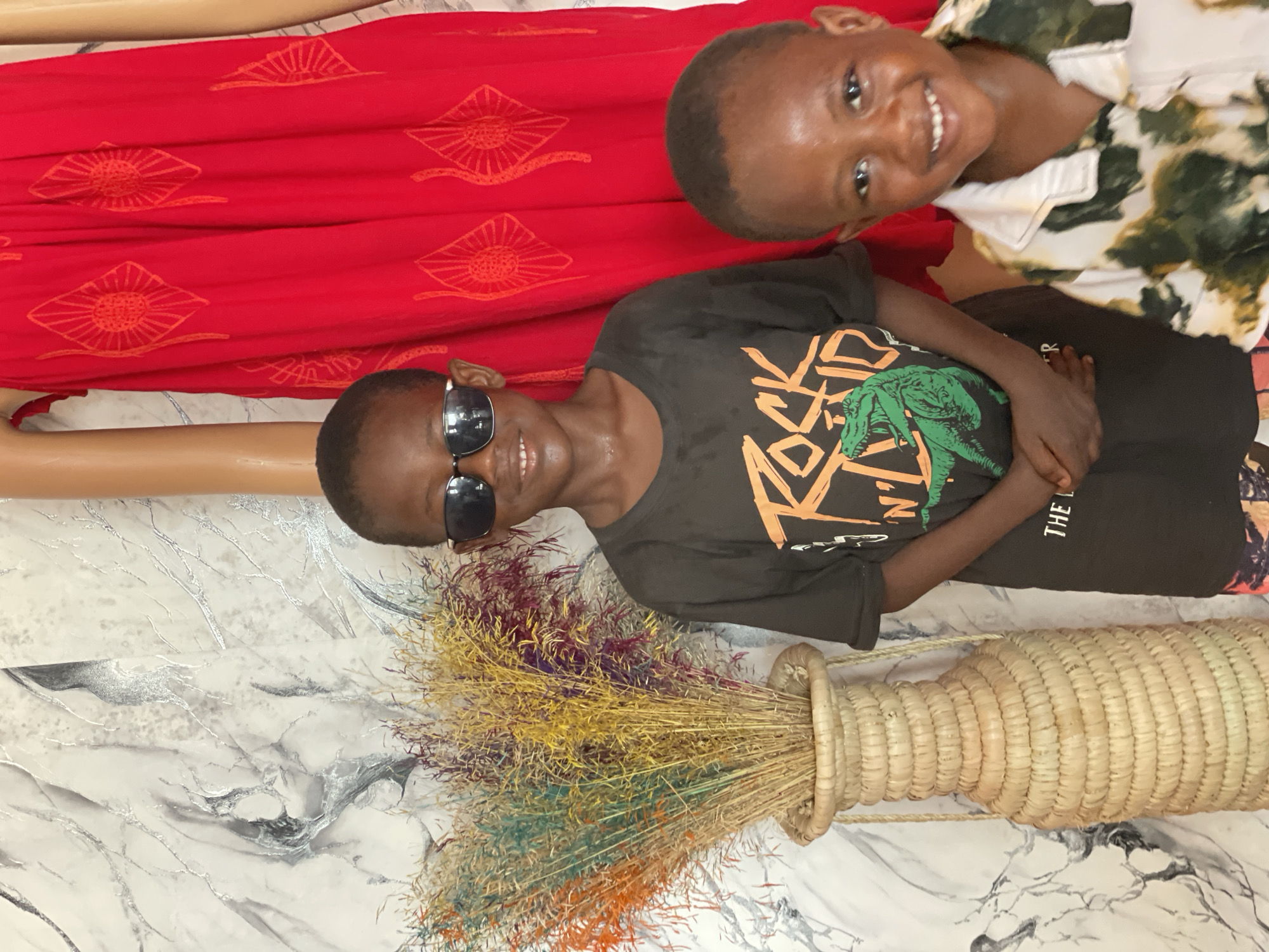 Two children are visible in an interior setting, possibly a shop or a home in Badagry, Nigeria. One child wears sunglasses and a black t-shirt with a green dinosaur design and lies near a decorative woven basket containing colorful dried grass. The second child is above and to the right, wearing a white and green patterned shirt. Behind them is a section of red fabric with repeating sun-like motifs, and a portion of a mannequin arm. The floor is a marble-patterned surface. The children are smiling and appear relaxed.